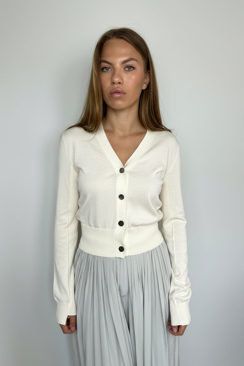A woman wearing a ivory button-front cardigan and a light gray pleated skirt, photographed against a plain light gray background.