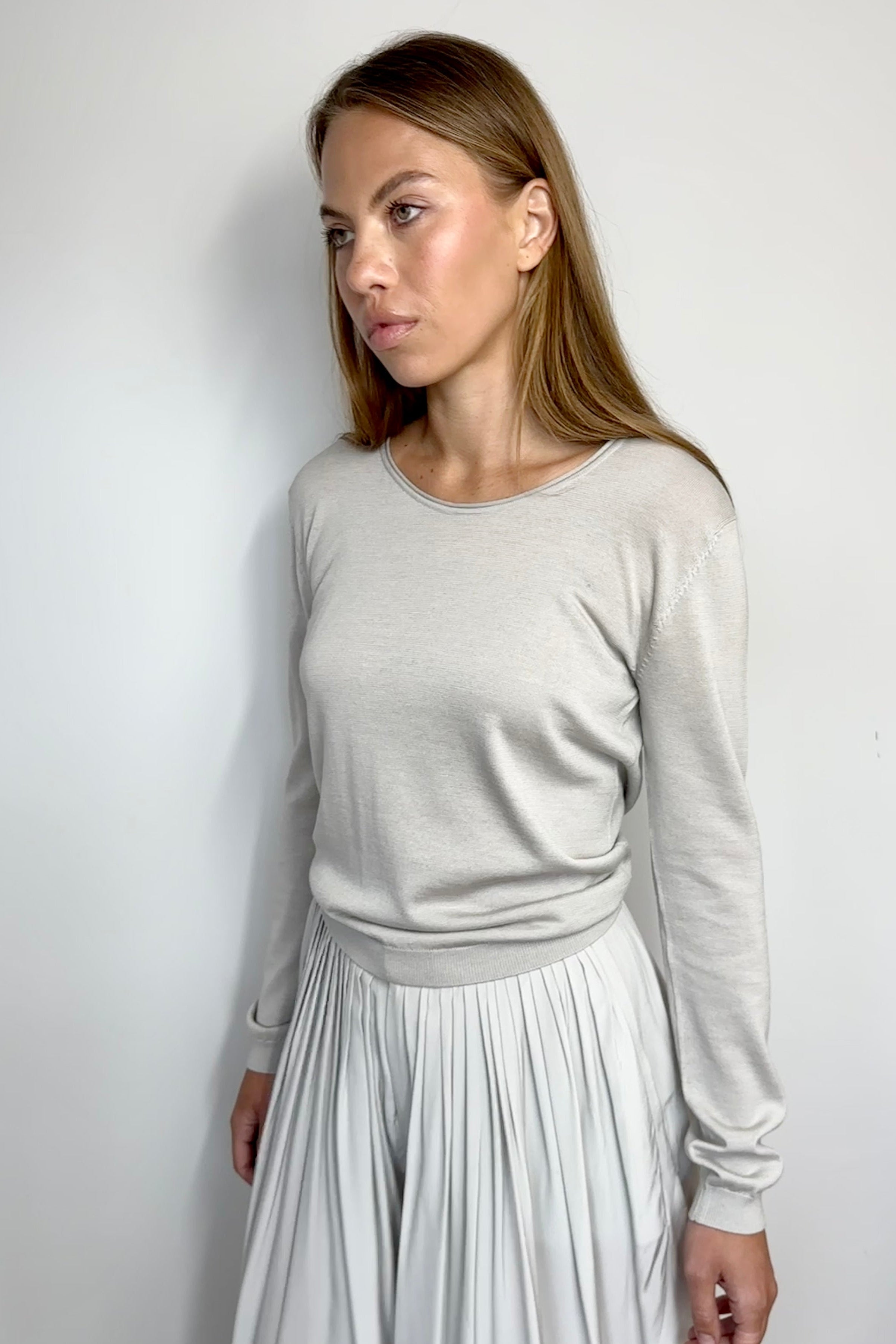 Woman wearing a light gray sweater and white pleated skirt against a plain background