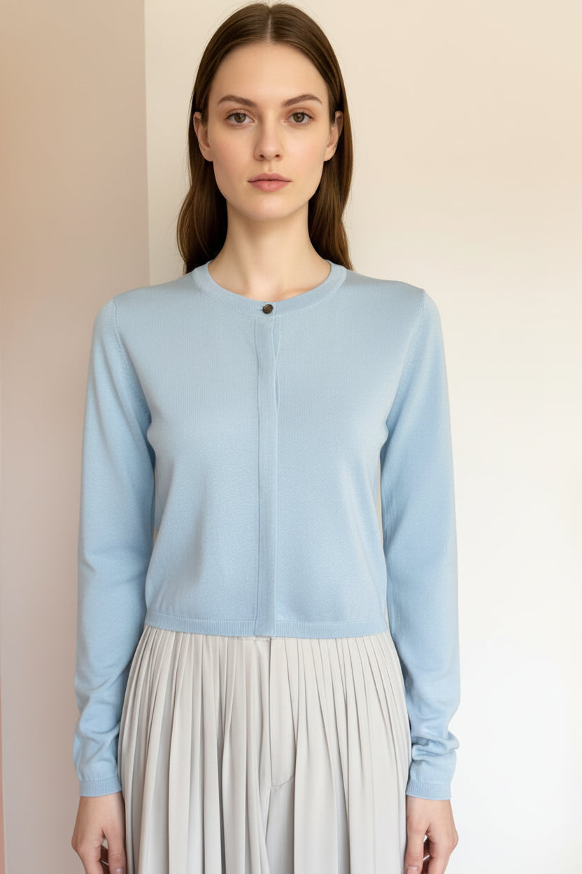 Woman wearing a light blue long-sleeve top and gray pleated skirt against a plain wall.