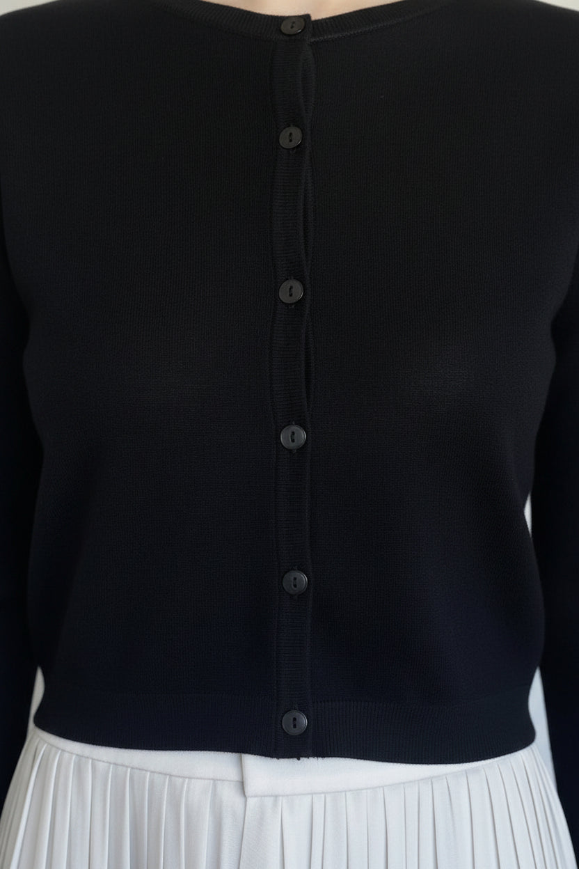 A black knit cardigan  with a button-down front on a model, wearing a white pleated skirt.