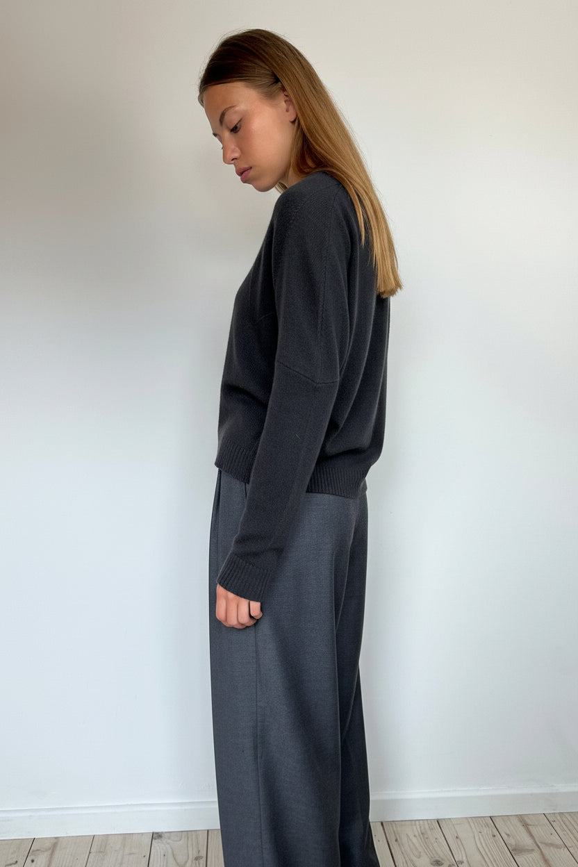 A woman stands in profile wearing a dark charcoal gray long-sleeve top and matching wide-leg trousers against a plain white wall and light wood floor.