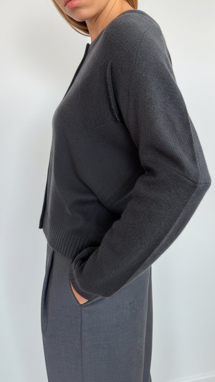 A woman wearing a dark charcoal gray ribbed knit sweater and matching gray skirt, shown from a side profile against a plain white wall.