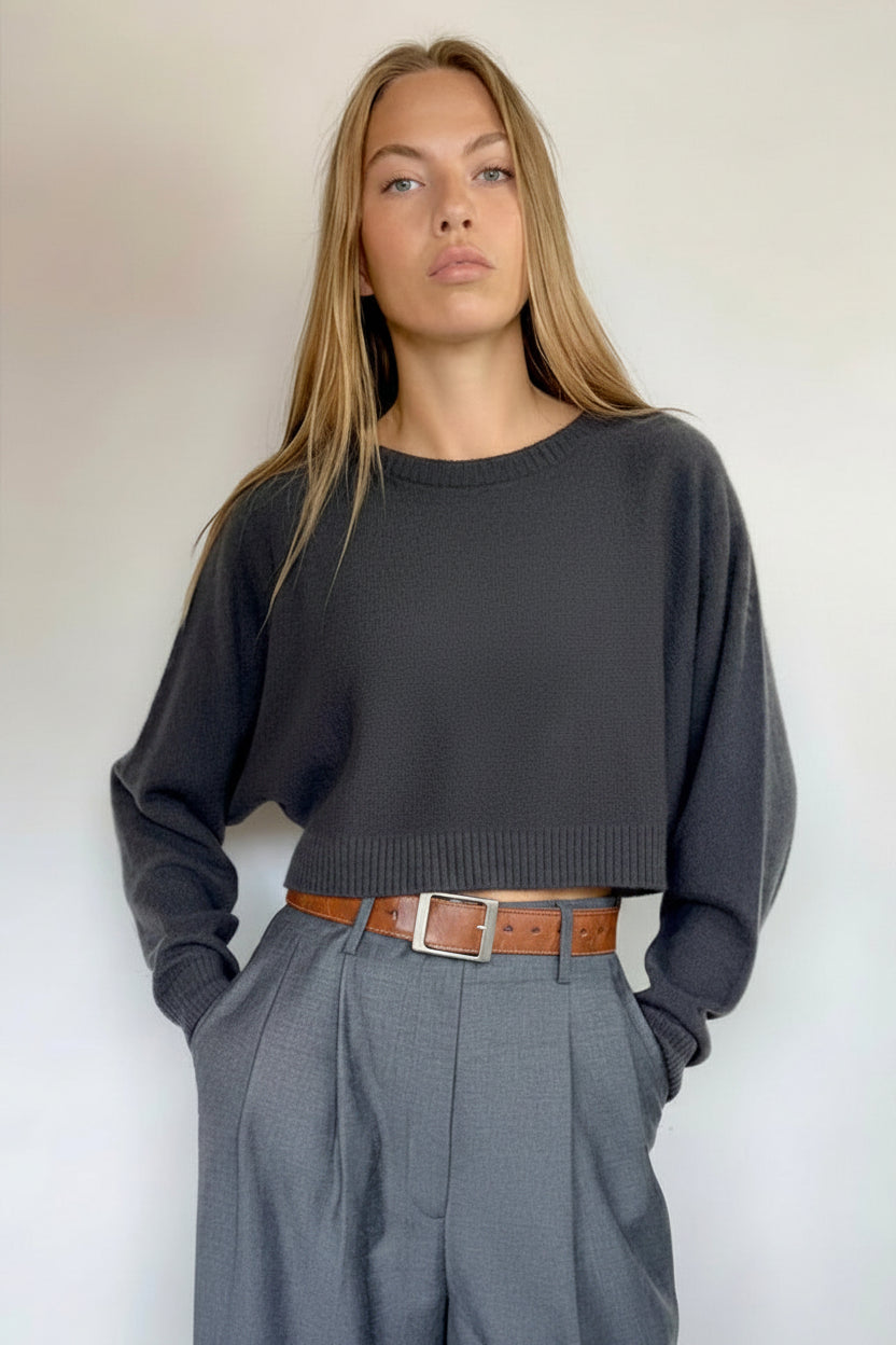 A woman modeling a dark charcoal gray cropped knit sweater and matching gray wide-leg trousers, styled with a brown leather belt, standing against a plain white wall.