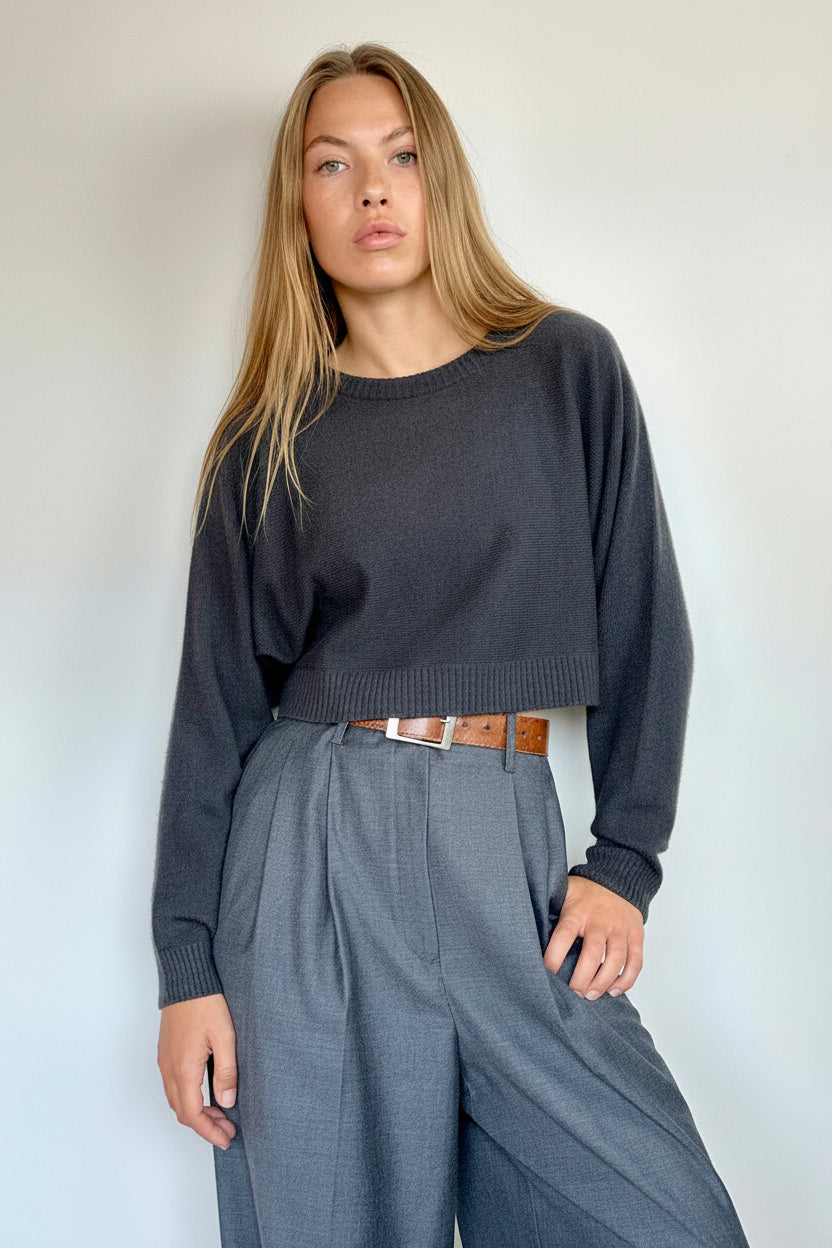A woman modeling a dark charcoal gray cropped knit sweater and slate blue wide-leg trousers with a brown leather belt, standing against a plain off-white wall.