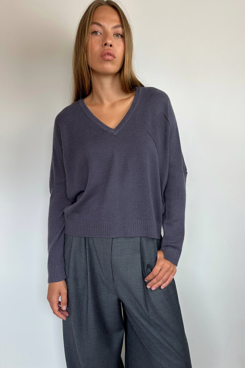 A woman modeling a dark slate purple V-neck knit sweater paired with dark gray wide-leg trousers, standing against a plain white wall.