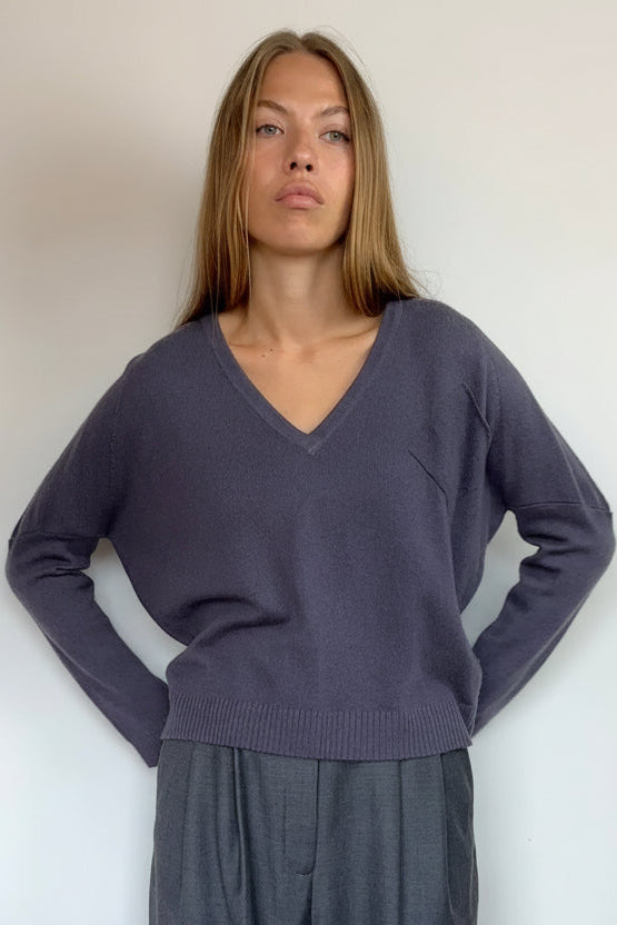 A woman modeling a slate purple v-neck knit sweater and dark gray wide-leg trousers against a plain white wall.