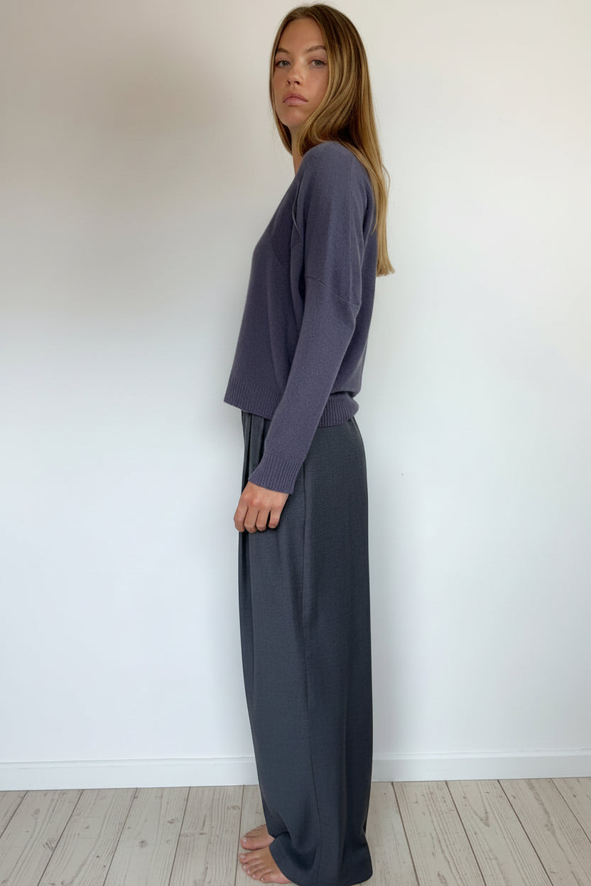 A woman wearing a slate purple knit long-sleeve crop sweater and dark gray wide-leg trousers, standing in profile against a plain white wall.