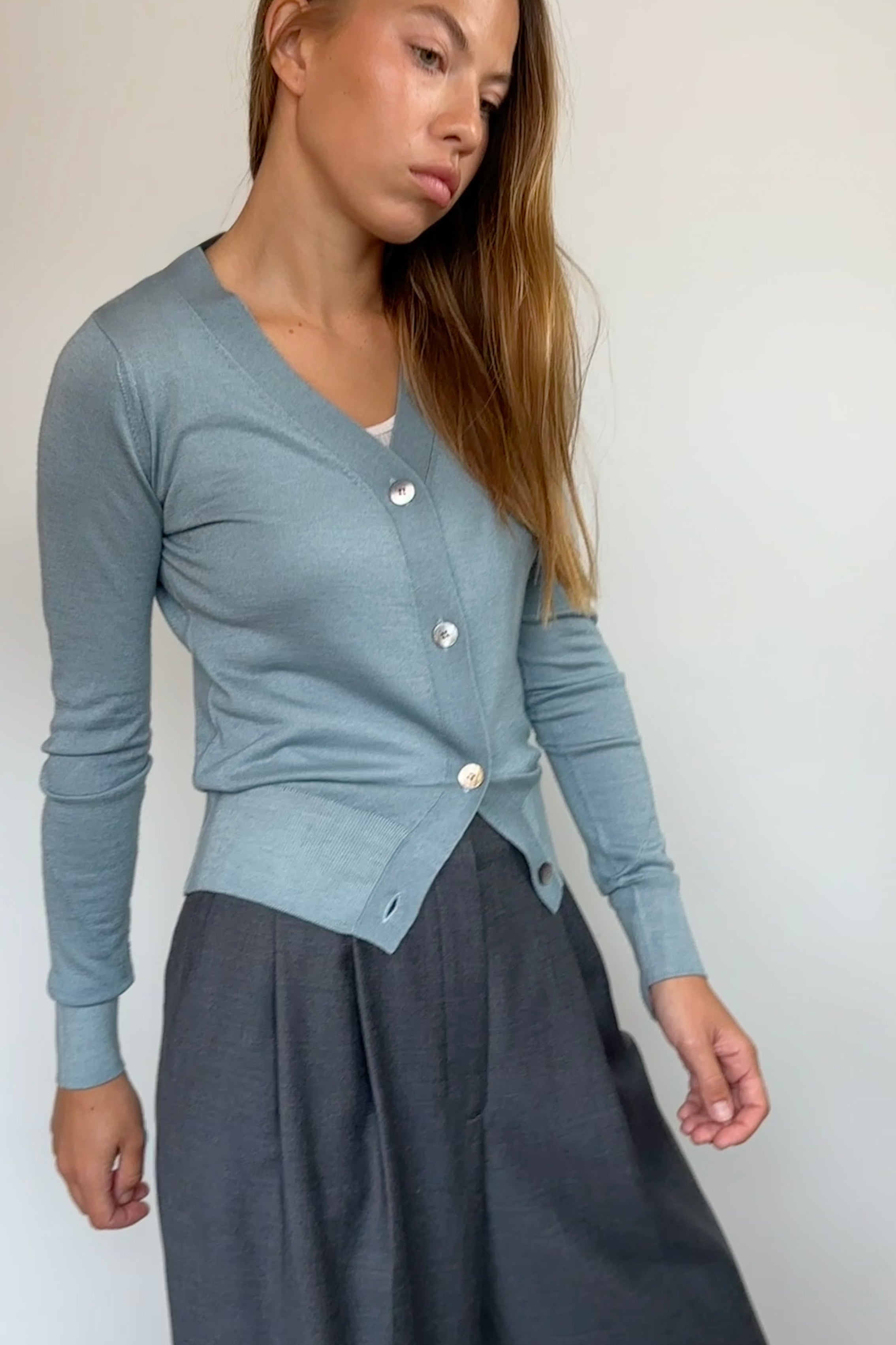 A woman wearing a light blue-gray long-sleeve cardigan with white buttons and a dark gray skirt, standing against a plain off-white wall.