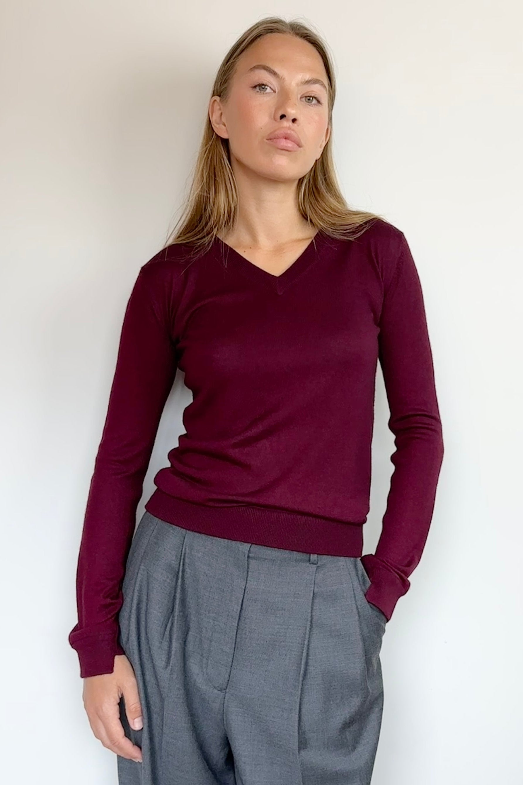 A woman modeling a long-sleeve V-neck knit sweater in deep burgundy, paired with gray wide-leg trousers, standing against a plain off-white wall.