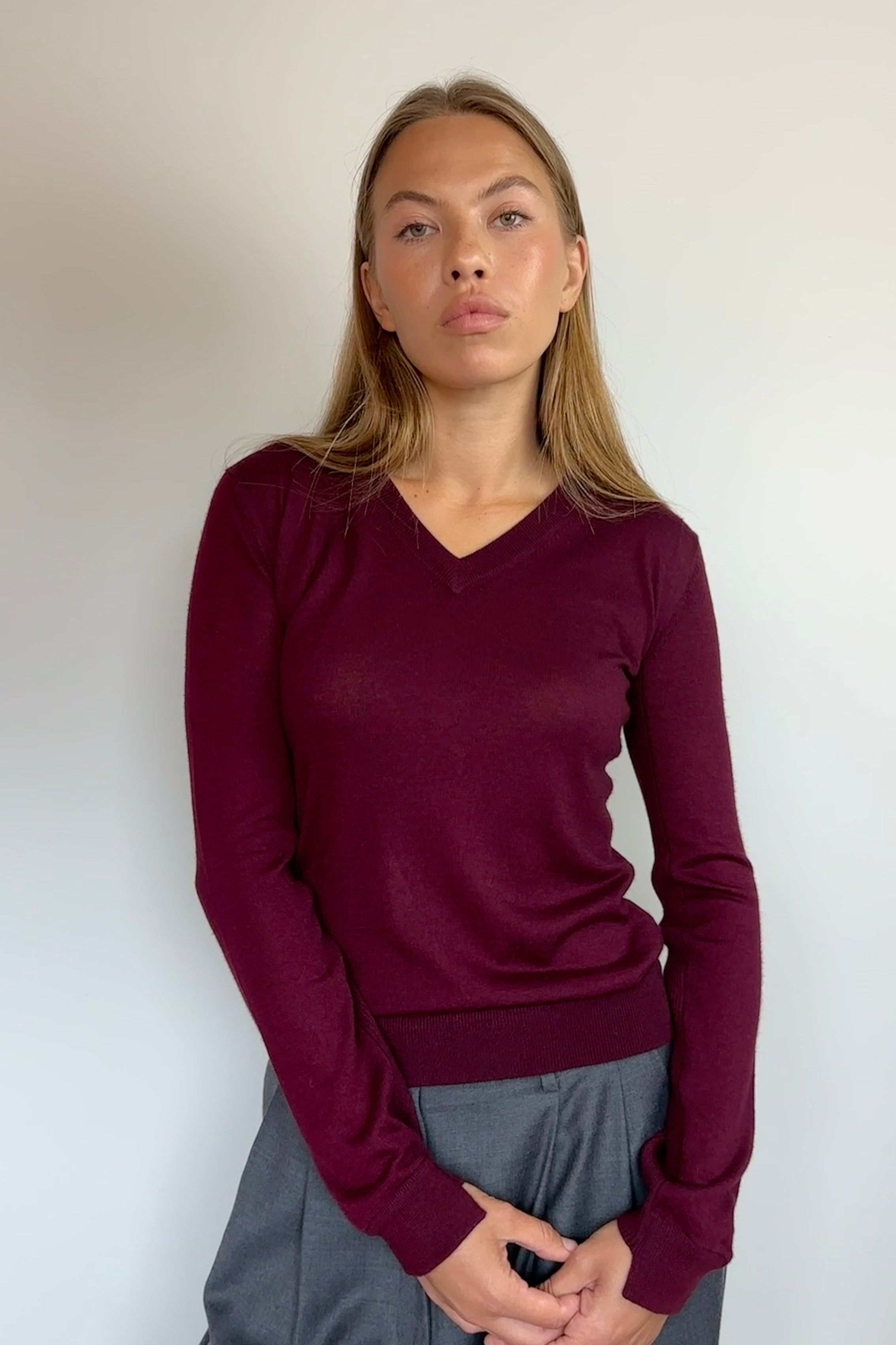 A woman wearing a long-sleeve v-neck knit sweater in deep burgundy, paired with gray trousers, standing against a plain off-white wall.