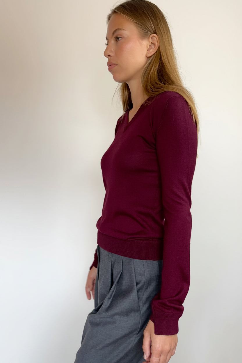 A woman in profile wearing a long-sleeve V-neck knit sweater in deep burgundy and gray trousers, standing against a plain off-white wall.
