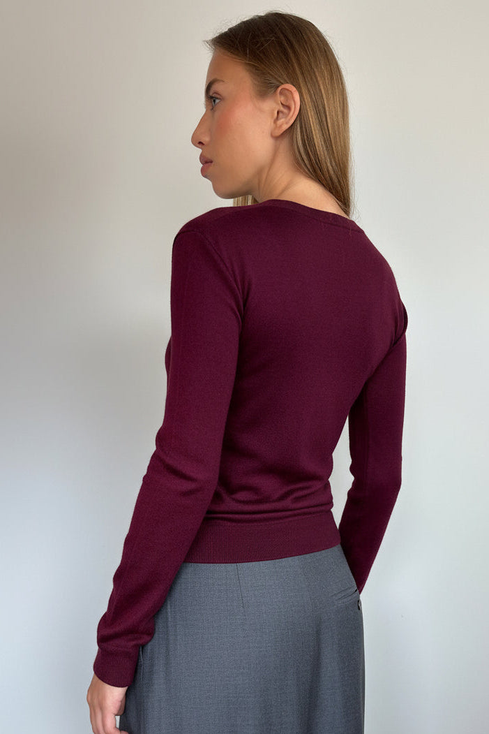 A woman shown from the back wearing a long-sleeve ribbed knit top in deep burgundy and a gray skirt, standing against a plain light gray wall.