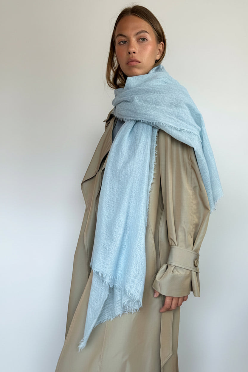 A woman wearing a light blue cashmere fringed scarf wrapped around her shoulders over a beige trench coat, standing against a plain off-white wall.
