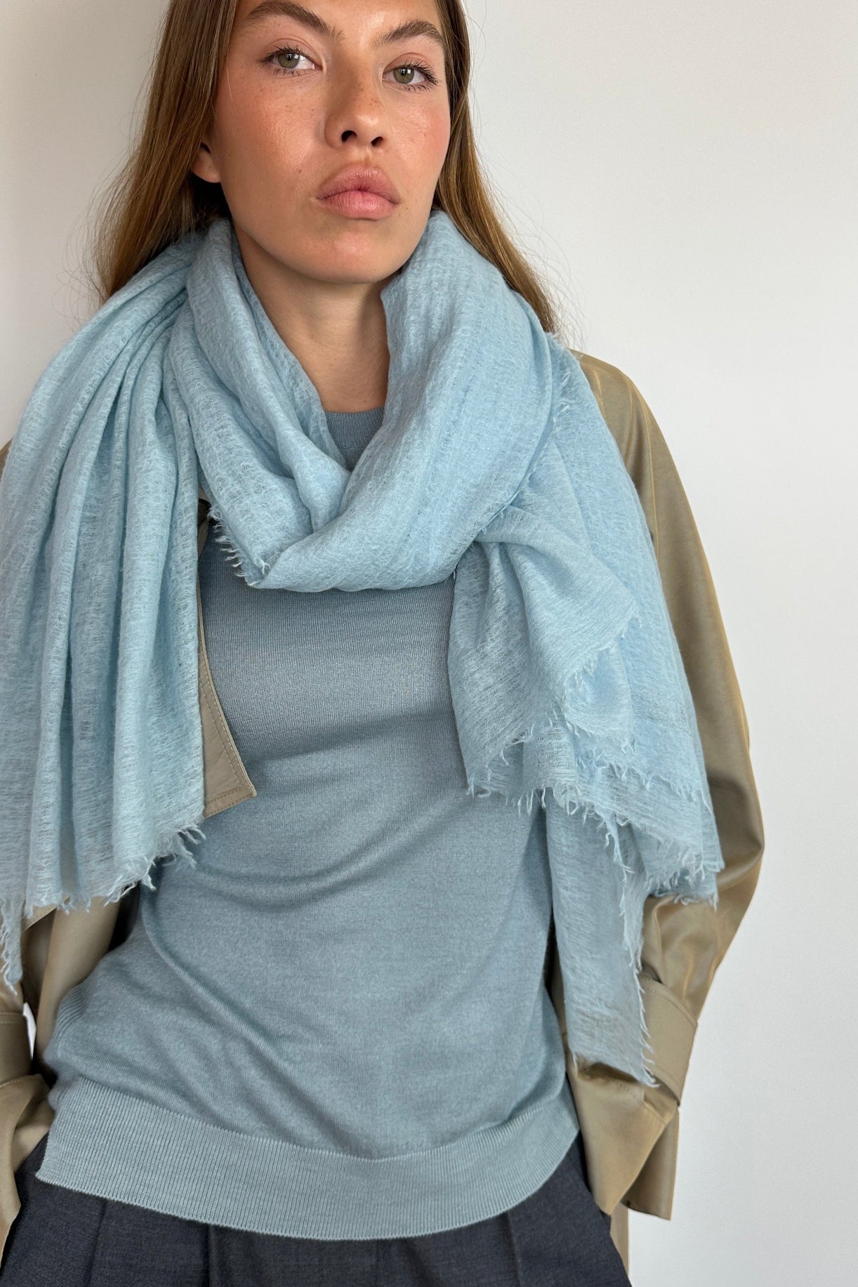 A woman wearing a light blue fringed scarf draped over her shoulders, paired with a gray top and a beige jacket, photographed against a plain light background.