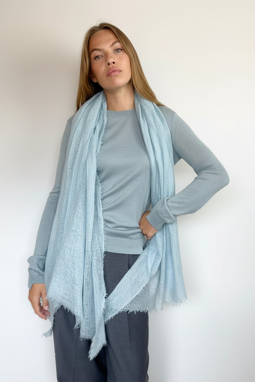 A woman modeling a light blue cashmere fringed scarf draped over a gray long-sleeve top and dark gray pants against a plain white wall.