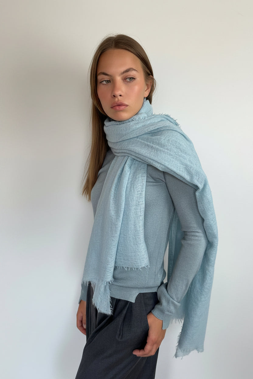 A woman modeling a light blue fringed scarf wrapped around her shoulders, paired with a gray long-sleeve top and dark gray pants, set against a plain off-white wall.