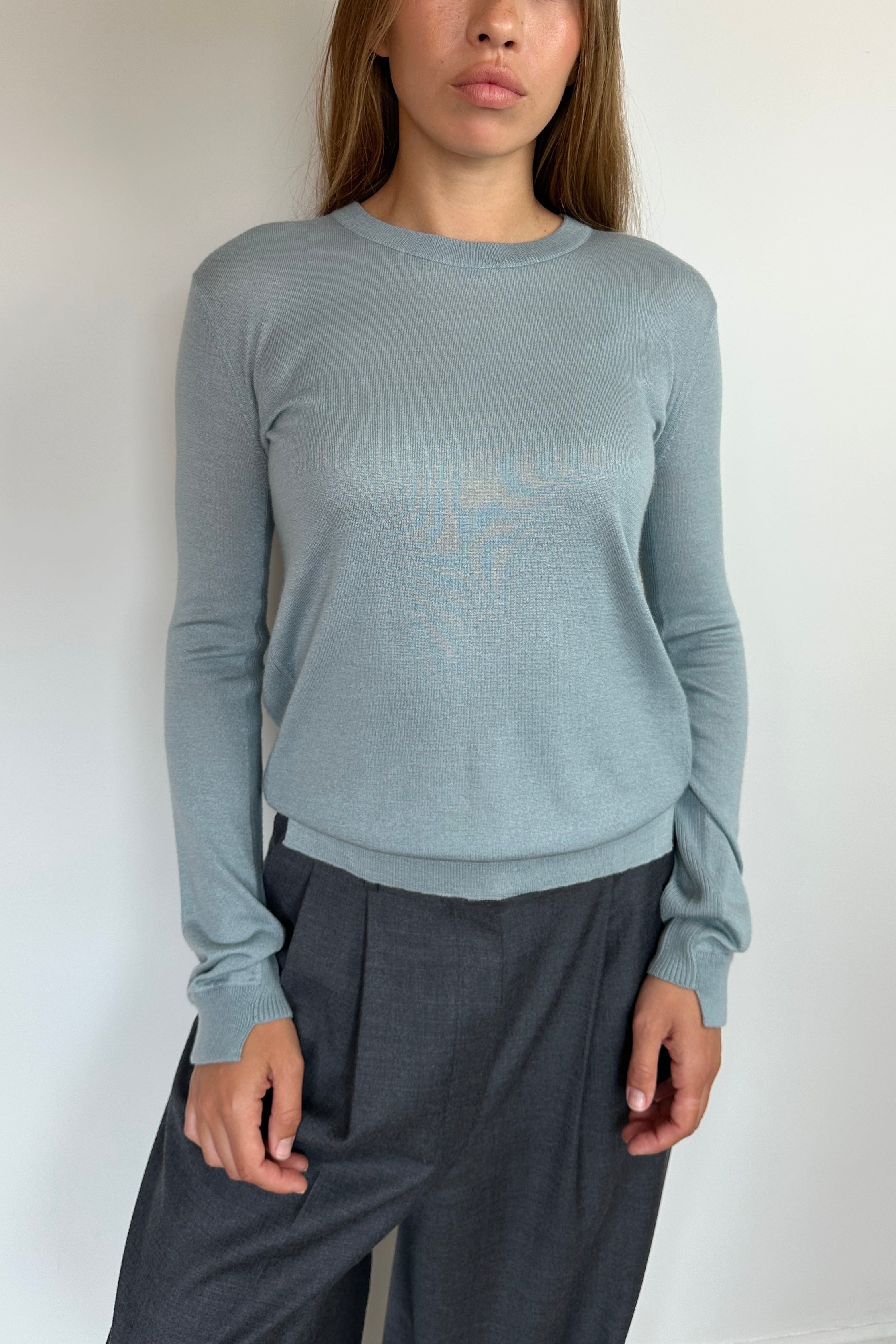 A woman wearing a light blue-gray long-sleeve knit sweater and dark gray wide-leg trousers, standing against a plain off-white wall.