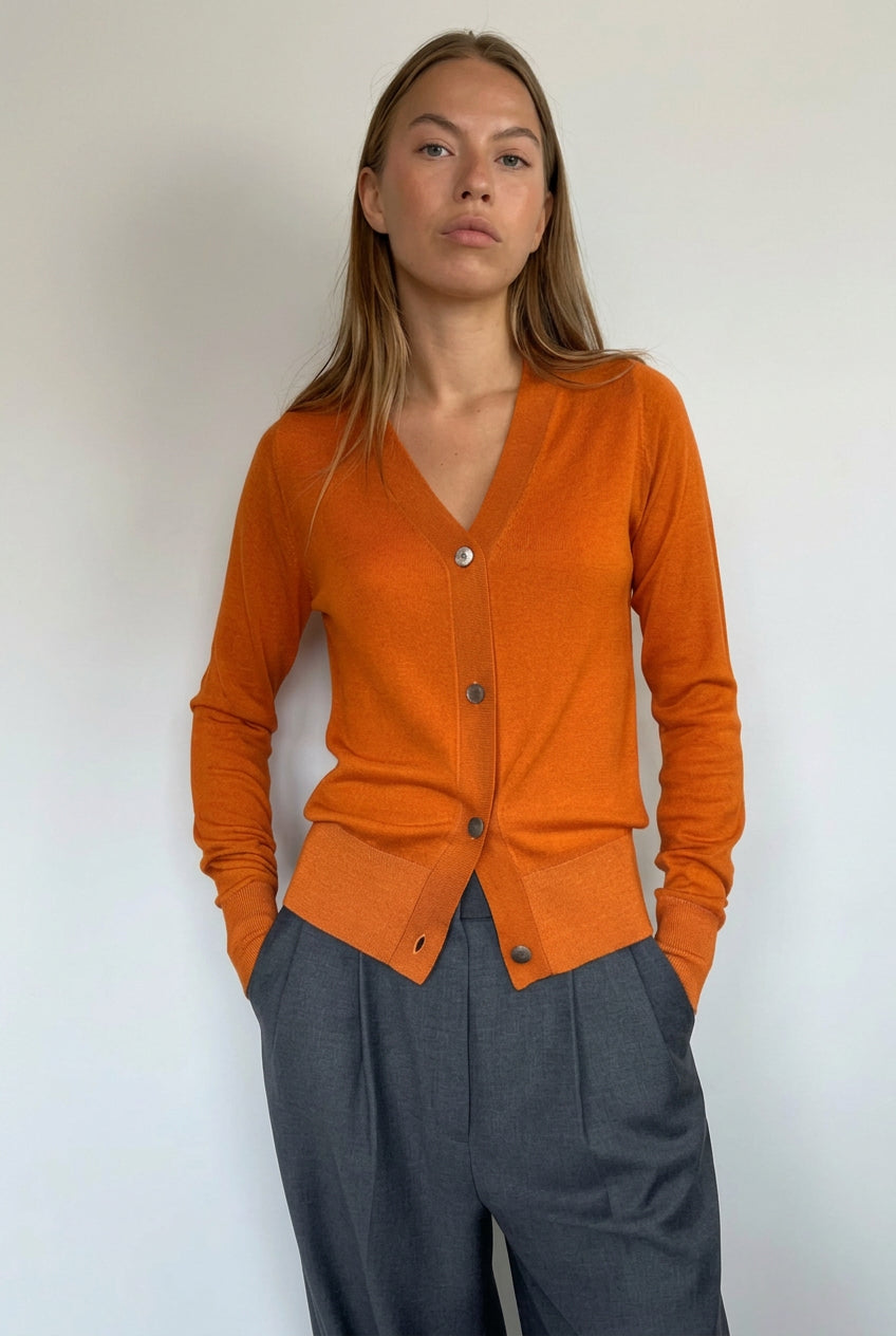 A woman modeling a bright orange long-sleeve button-front cardigan with a V-neck, paired with dark gray wide-leg pants, standing against a plain light gray wall.