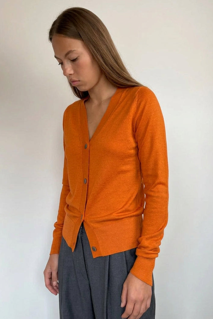A woman wearing a vibrant orange long-sleeve button-up cardigan and dark gray wide-leg trousers, standing against a plain off-white wall.
