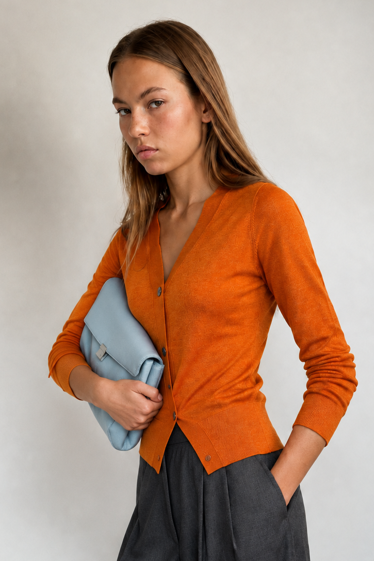 A woman models a vibrant orange knit cardigan with a V-neck and button closure, styled with dark gray wide-leg trousers and a light blue clutch, set against a neutral light gray background.