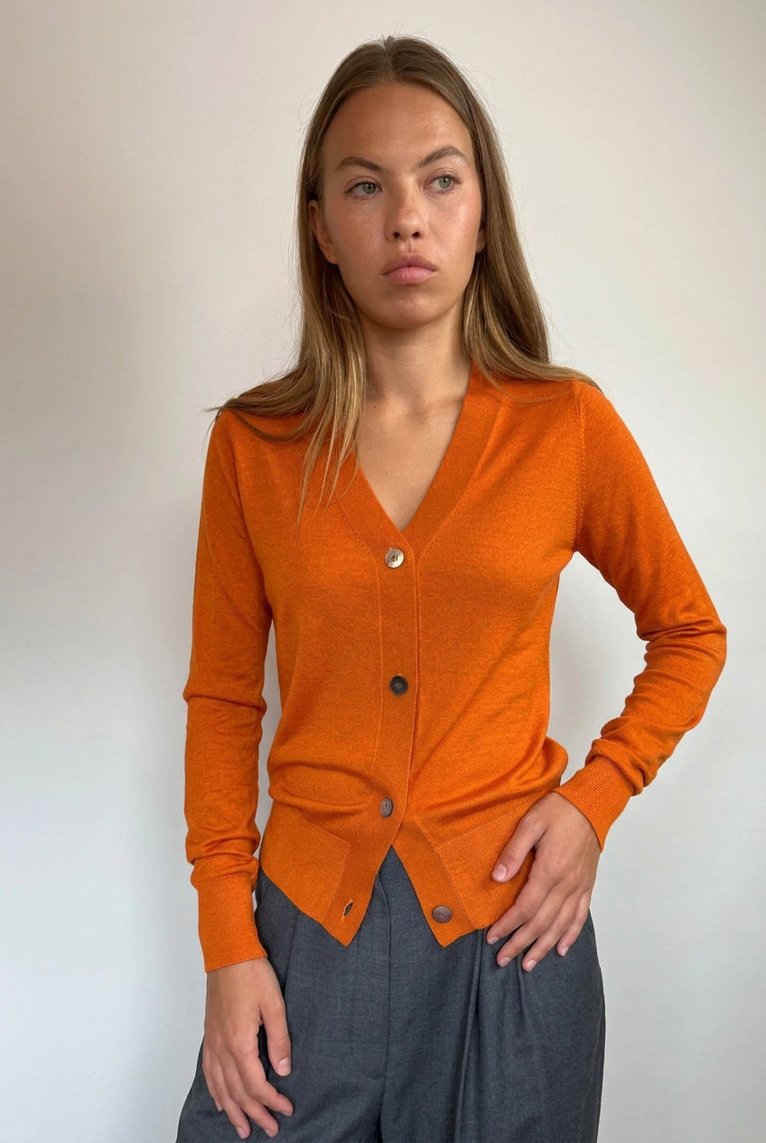 A woman modeling a rust-orange long-sleeve button-up cardigan sweater paired with dark gray wide-leg pants, standing against a plain off-white wall.