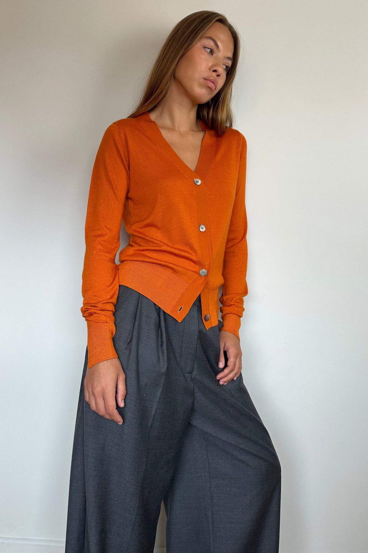 A woman wearing a bright orange button-front cardigan with long sleeves and dark gray wide-leg trousers, standing against a plain off-white wall.