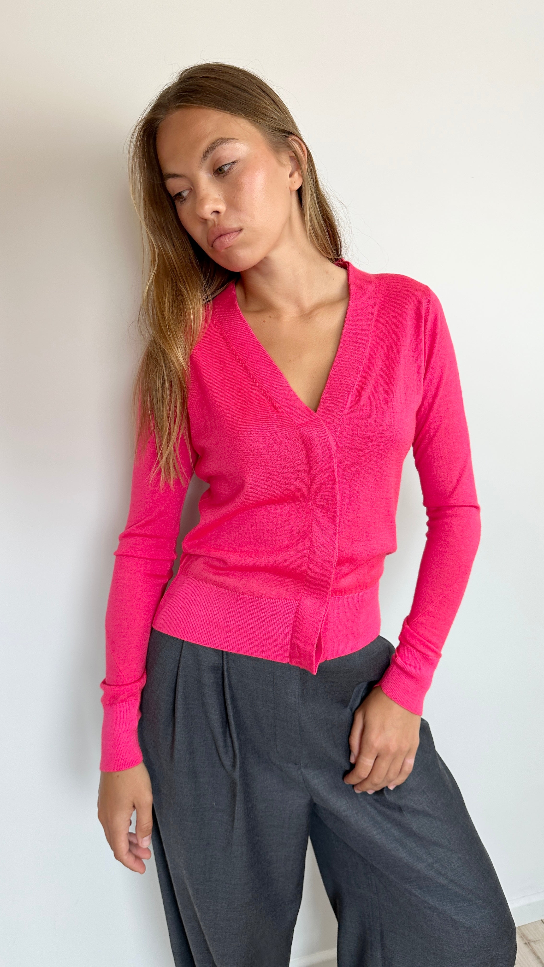 A woman wearing a bright pink long-sleeve V-neck cardigan and dark gray wide-leg trousers, standing against a plain white wall.
