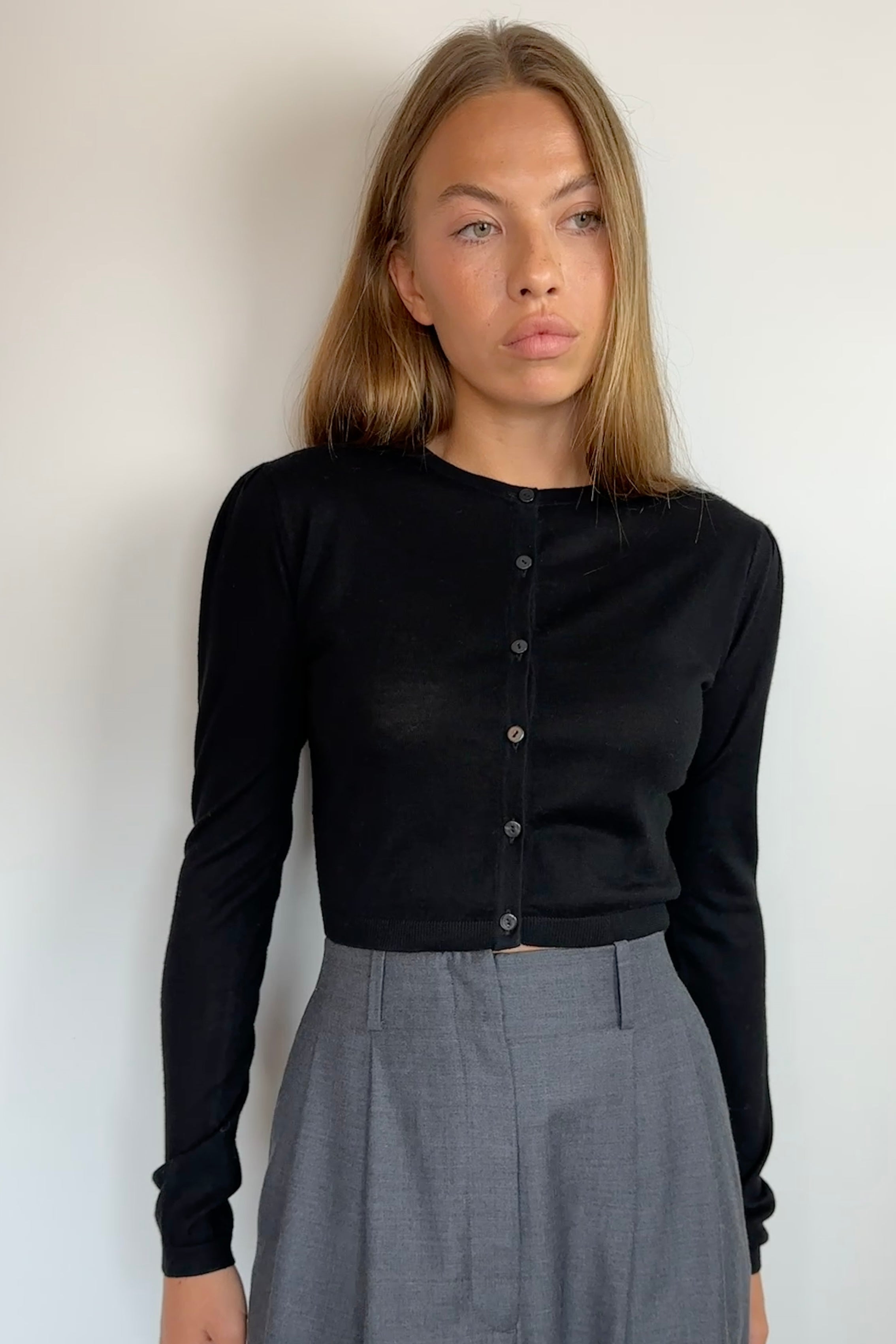 A woman wearing a black long-sleeve button-front top and gray trousers, standing against a plain light background.