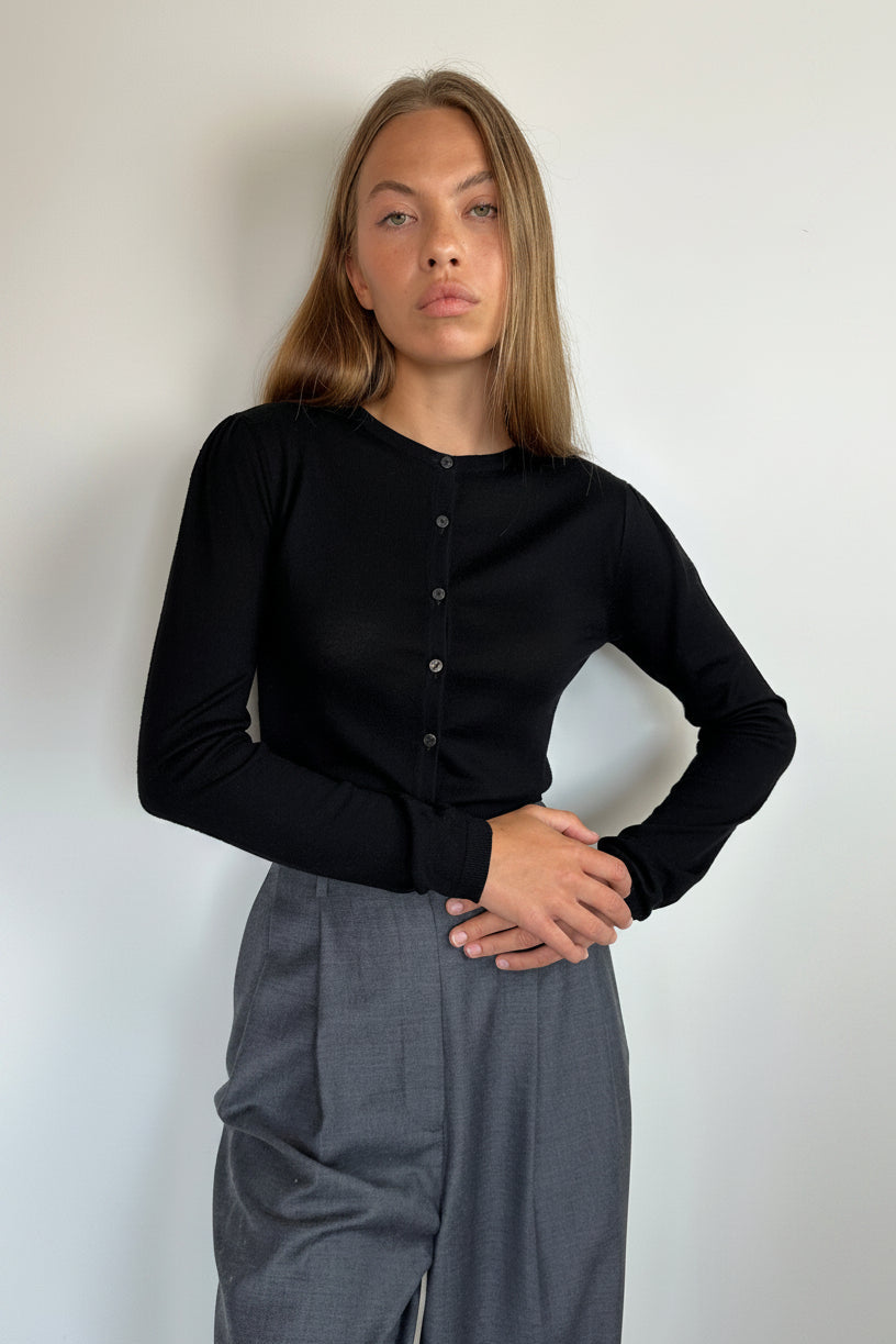 A woman modeling a black long-sleeve button-front top with gray wide-leg trousers, standing against a plain light gray wall.