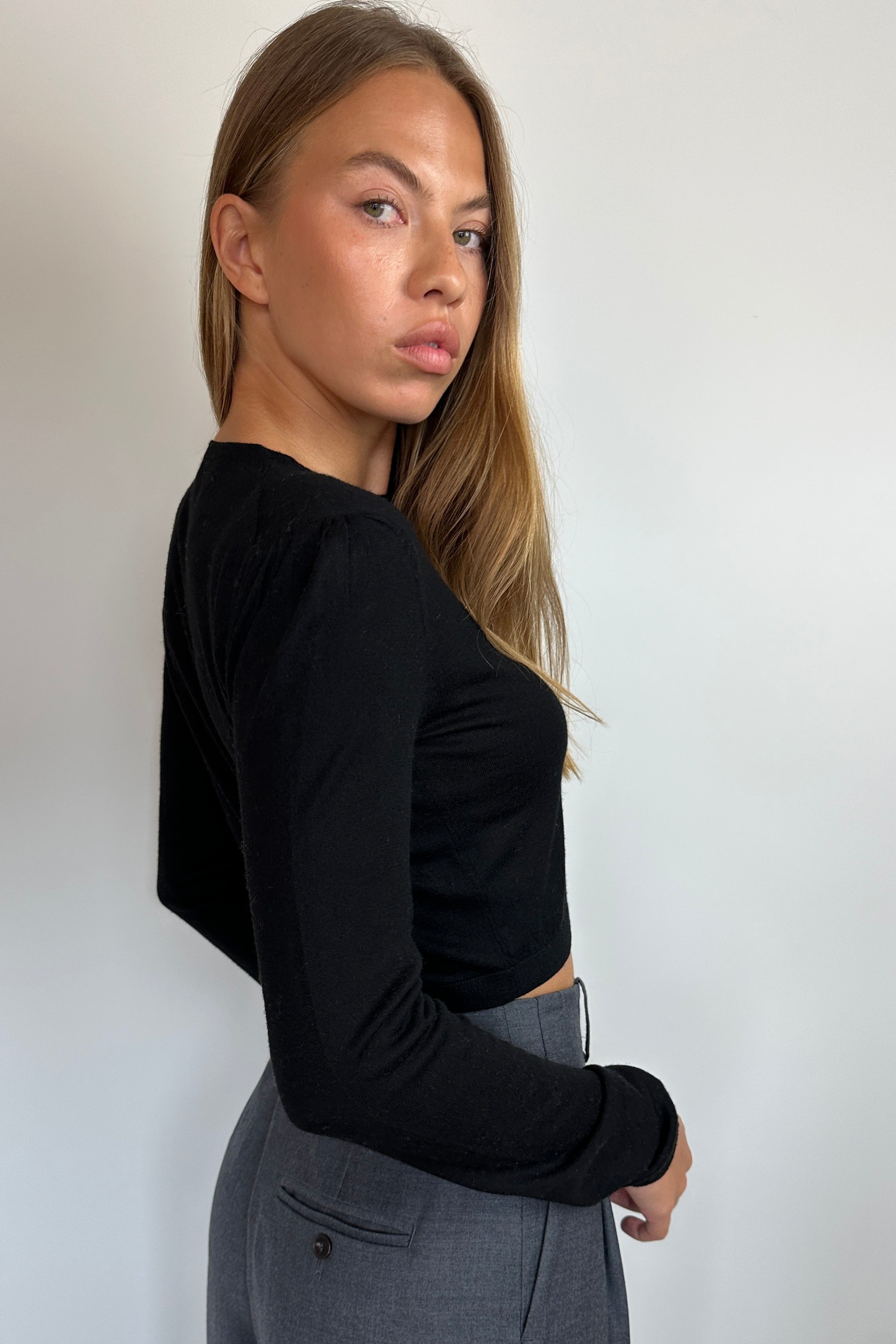 A woman wearing a black long-sleeve crop top and gray trousers, photographed from the side against a plain light gray wall.