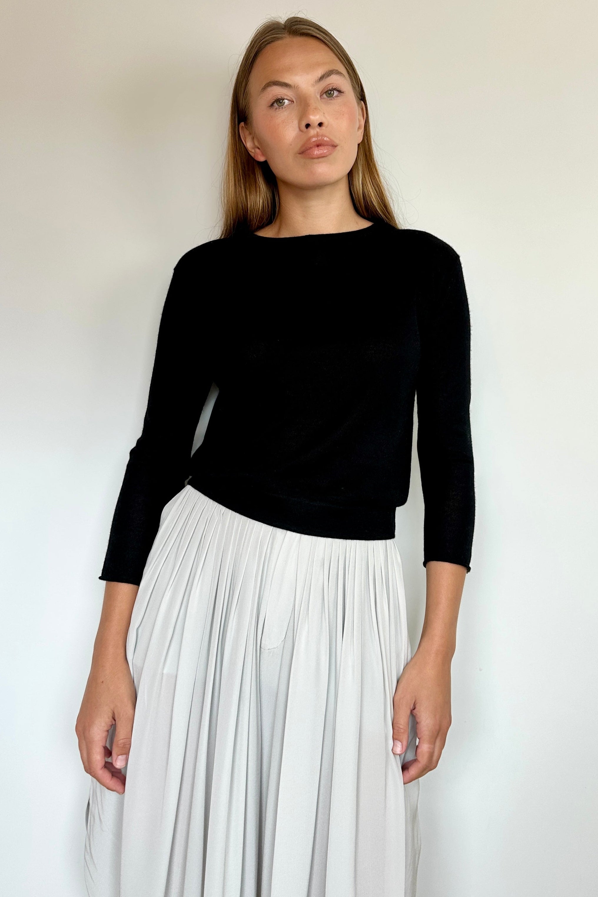 A woman modeling a black long-sleeve knit top paired with a high-waisted, pleated white skirt, standing against a plain off-white wall.
