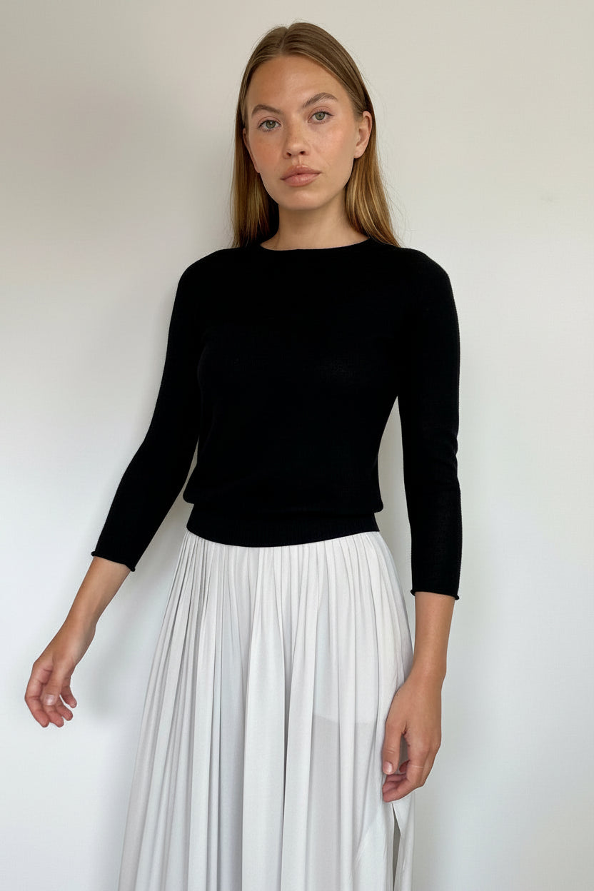 A woman modeling a black long-sleeve top paired with a high-waisted white pleated skirt, standing against a plain light gray wall.