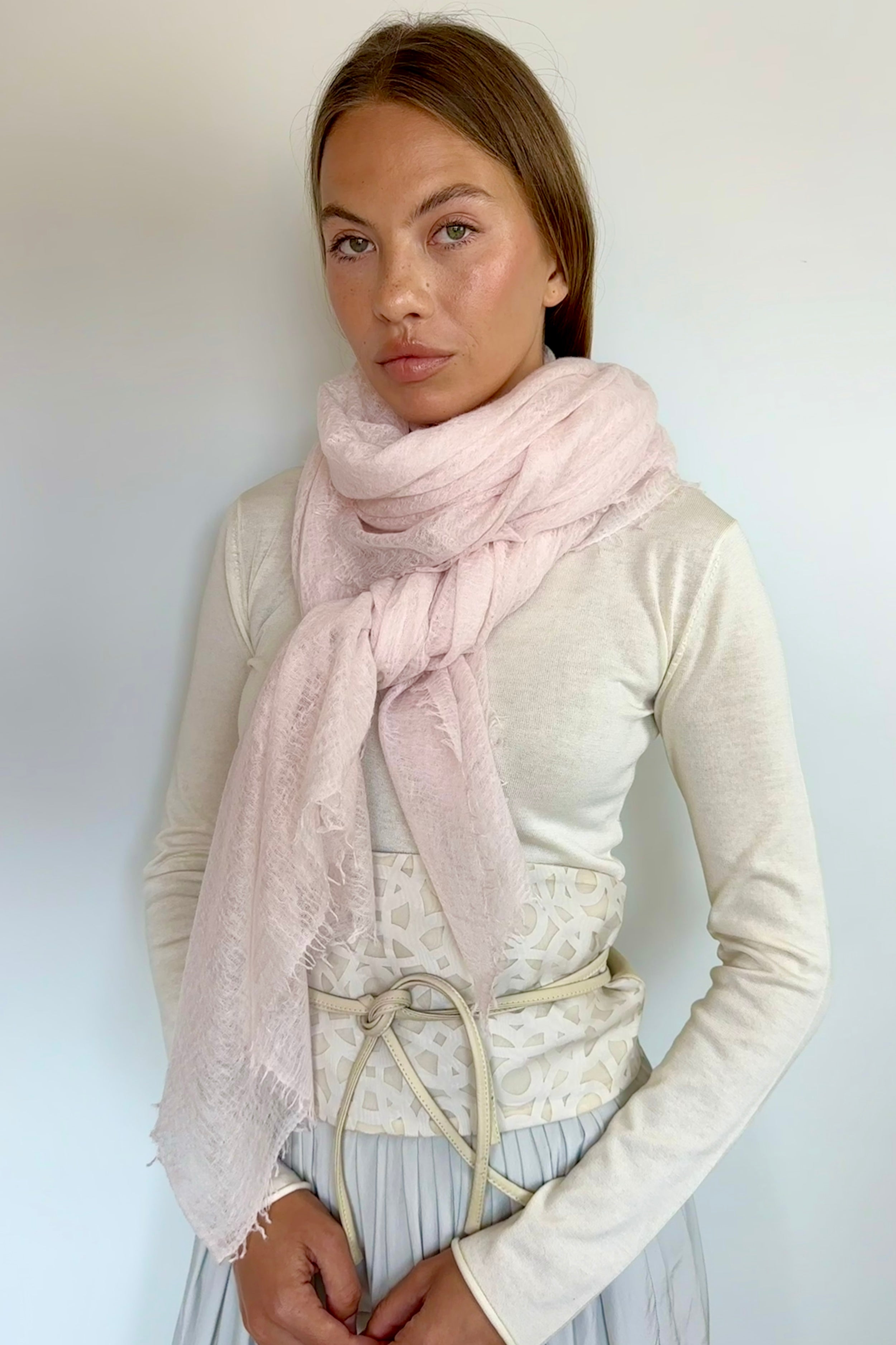 A woman wearing a light pink fringed scarf wrapped around her neck, paired with a cream-colored long-sleeve top and a pleated skirt, standing against a plain light gray wall.