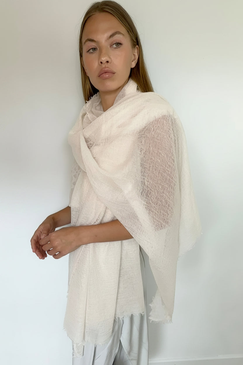 A woman modeling a lightweight, semi-sheer off-white shawl or wrap with a textured knit pattern, styled over a light gray dress against a plain white wall.
