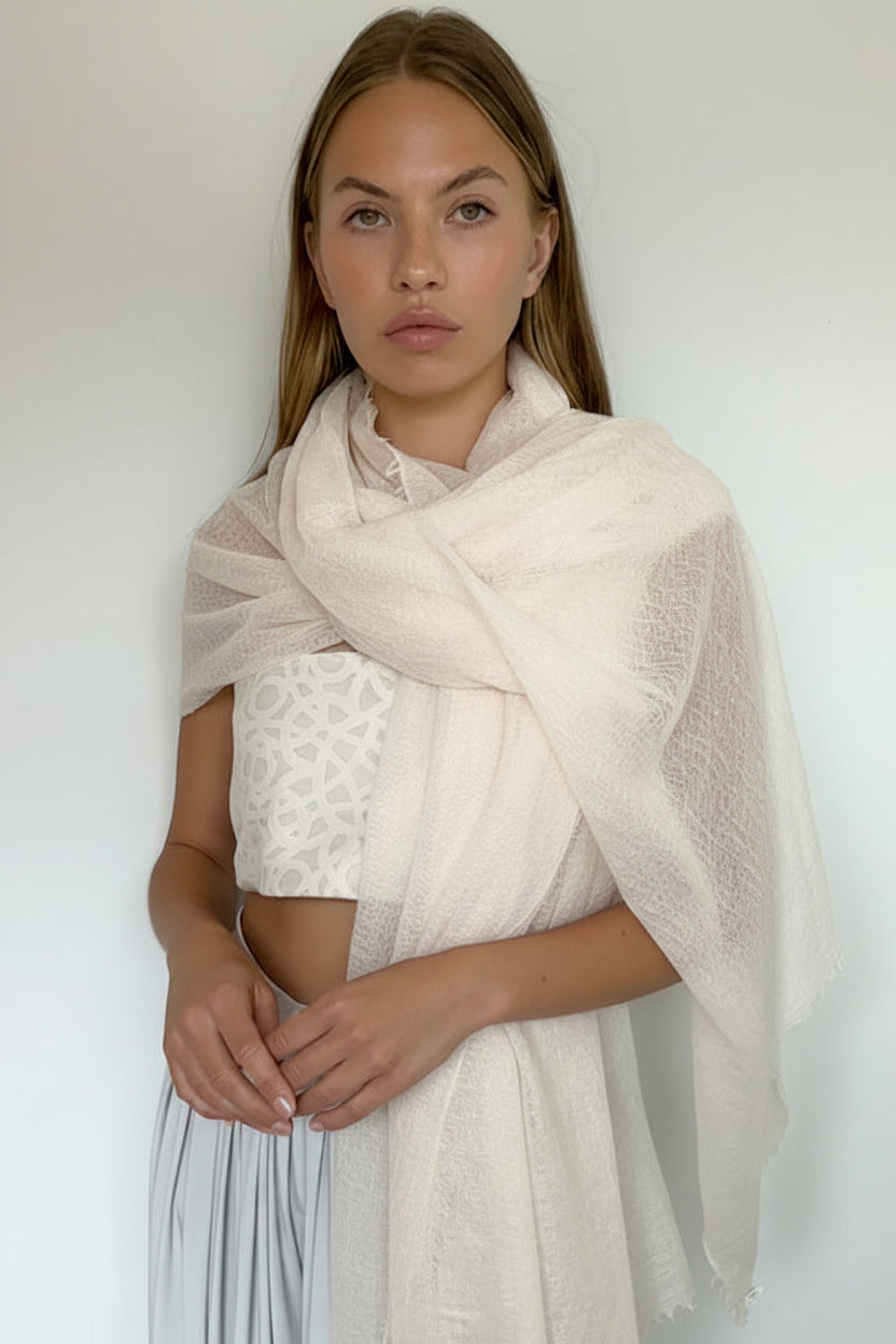 A woman modeling a light cream/off-white sheer lace shawl draped over her shoulders, paired with a white lace crop top and white pleated skirt, standing against a plain white wall.