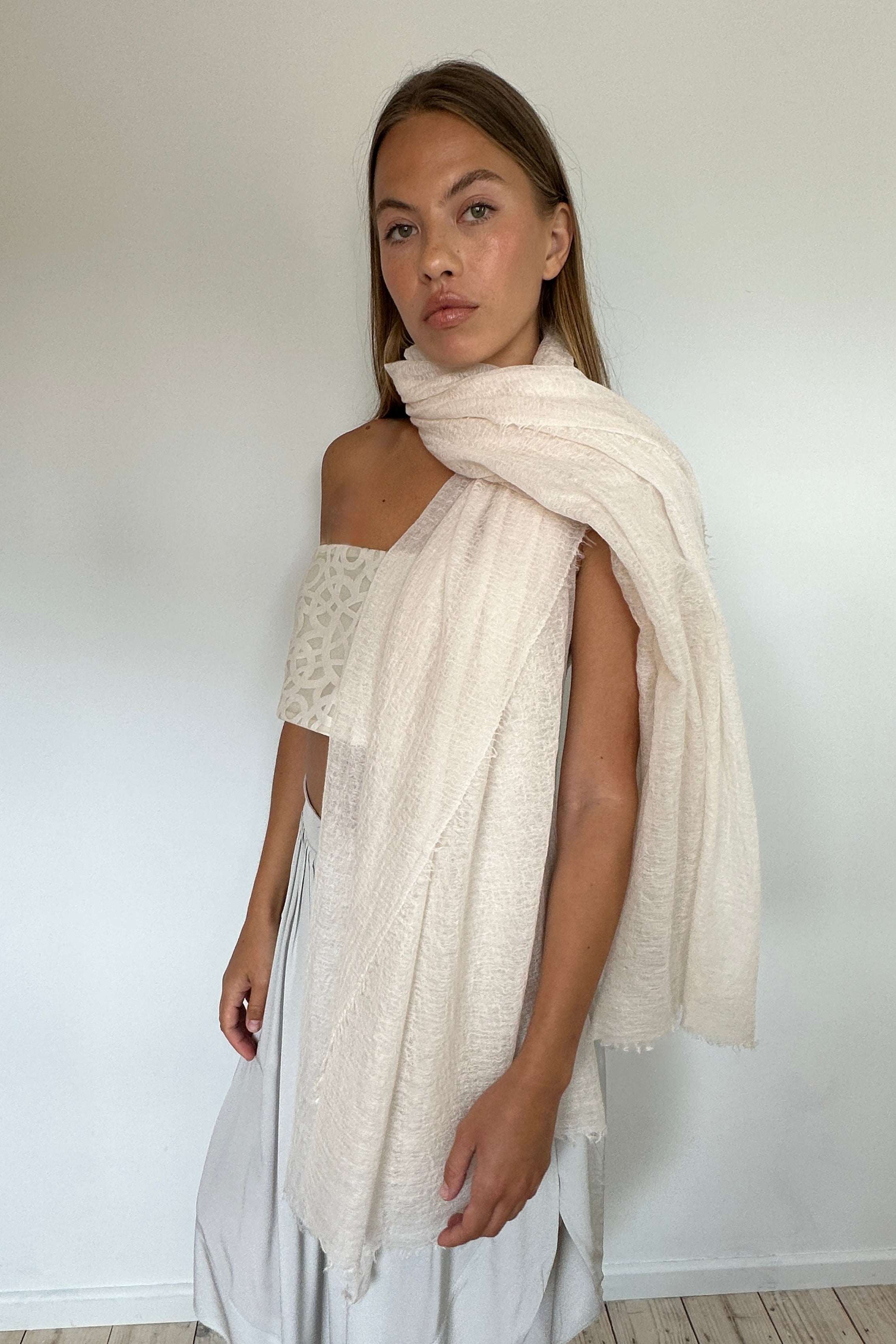 A woman modeling a light cream/off-white textured knit shawl draped over her shoulders, paired with a white lace off-the-shoulder top and white skirt, standing against a plain white wall and light wood floor.