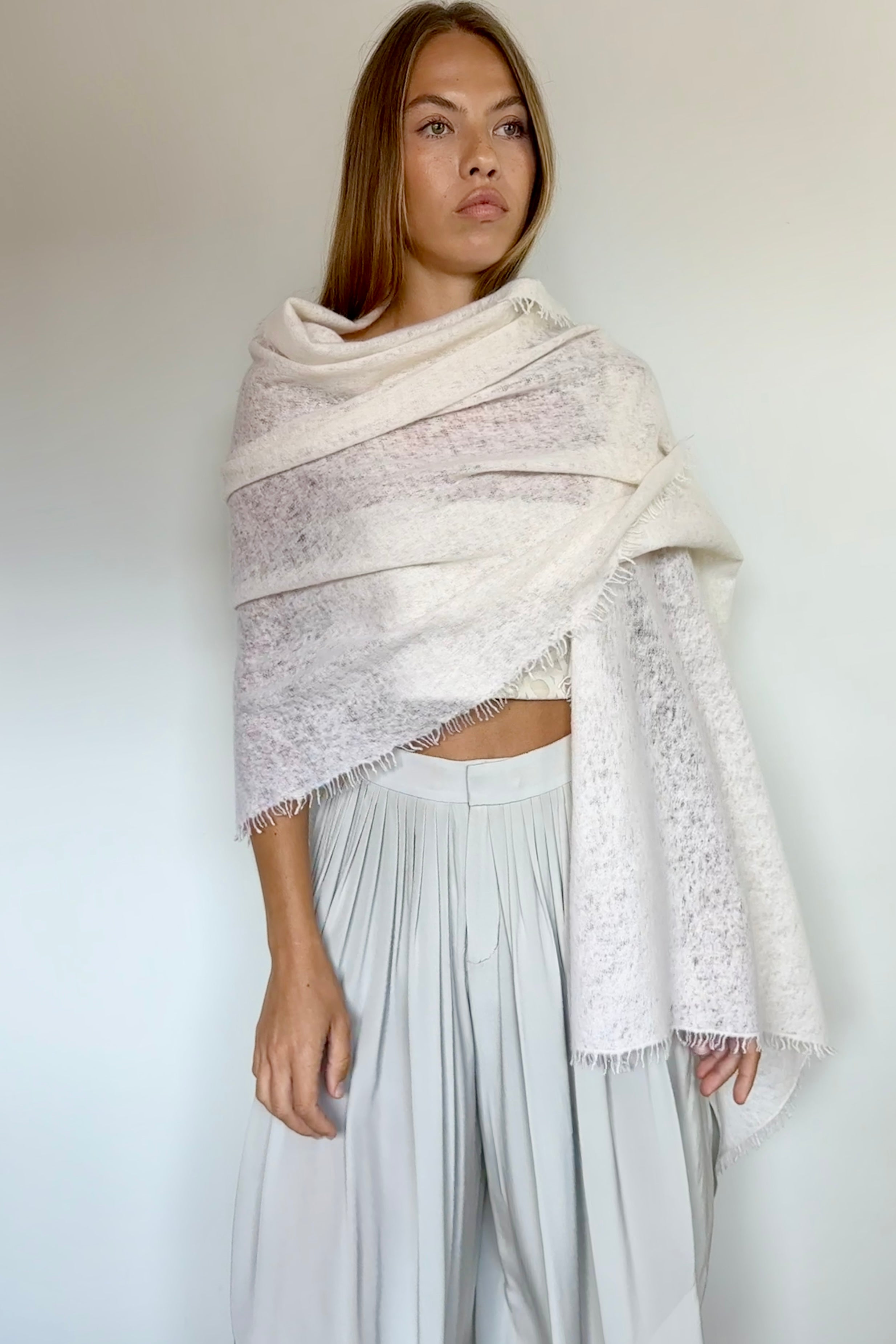 A woman wearing a large, textured cream-colored shawl with fringed edges draped over her shoulders, paired with light gray wide-leg trousers, standing against a plain off-white wall.