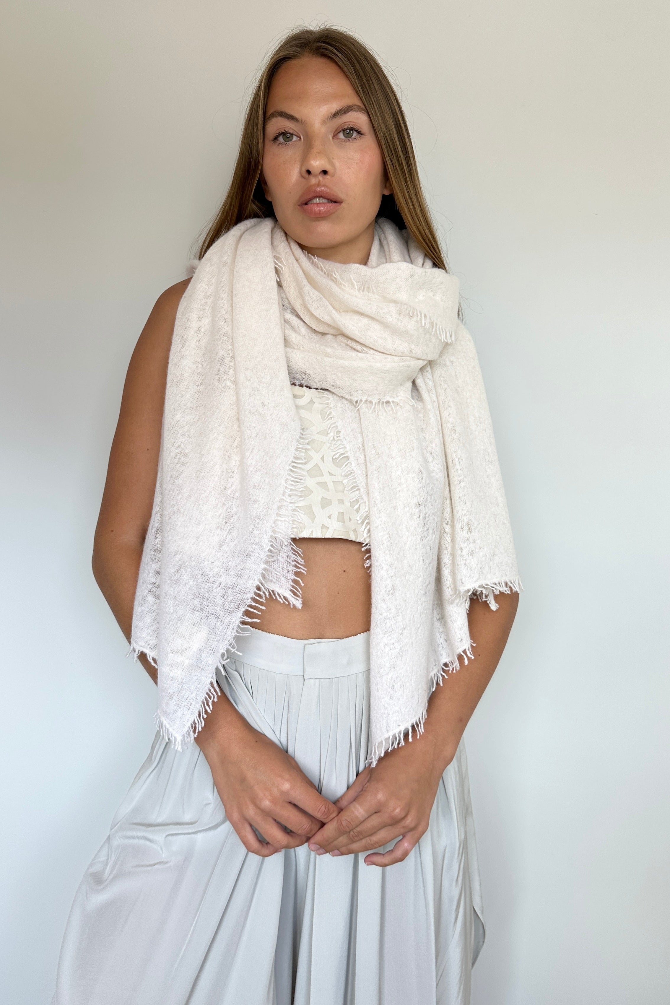 A woman modeling a cream-colored textured fringed scarf wrapped around her shoulders, paired with a matching light gray crop top and wide-leg pants, set against a plain light gray wall.