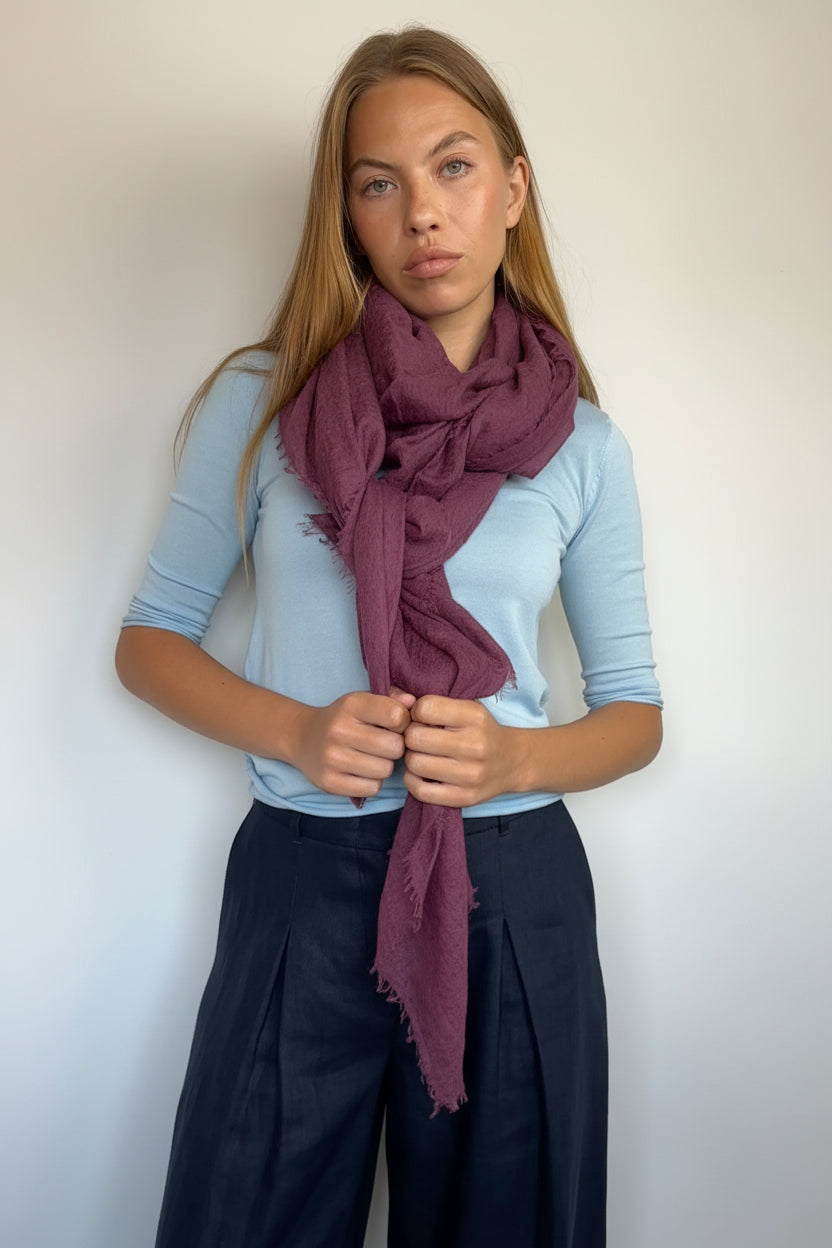 A woman modeling a dusty plum purple scarf tied around her neck, paired with a light blue long-sleeve top and dark pleated trousers, set against a plain off-white wall.