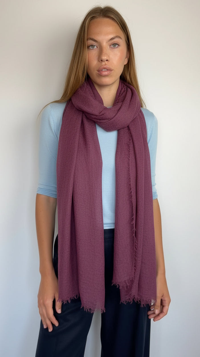 A woman modeling a lightweight, semi-sheer scarf in a dusty plum purple color, styled around her neck over a light blue t-shirt and dark pants.