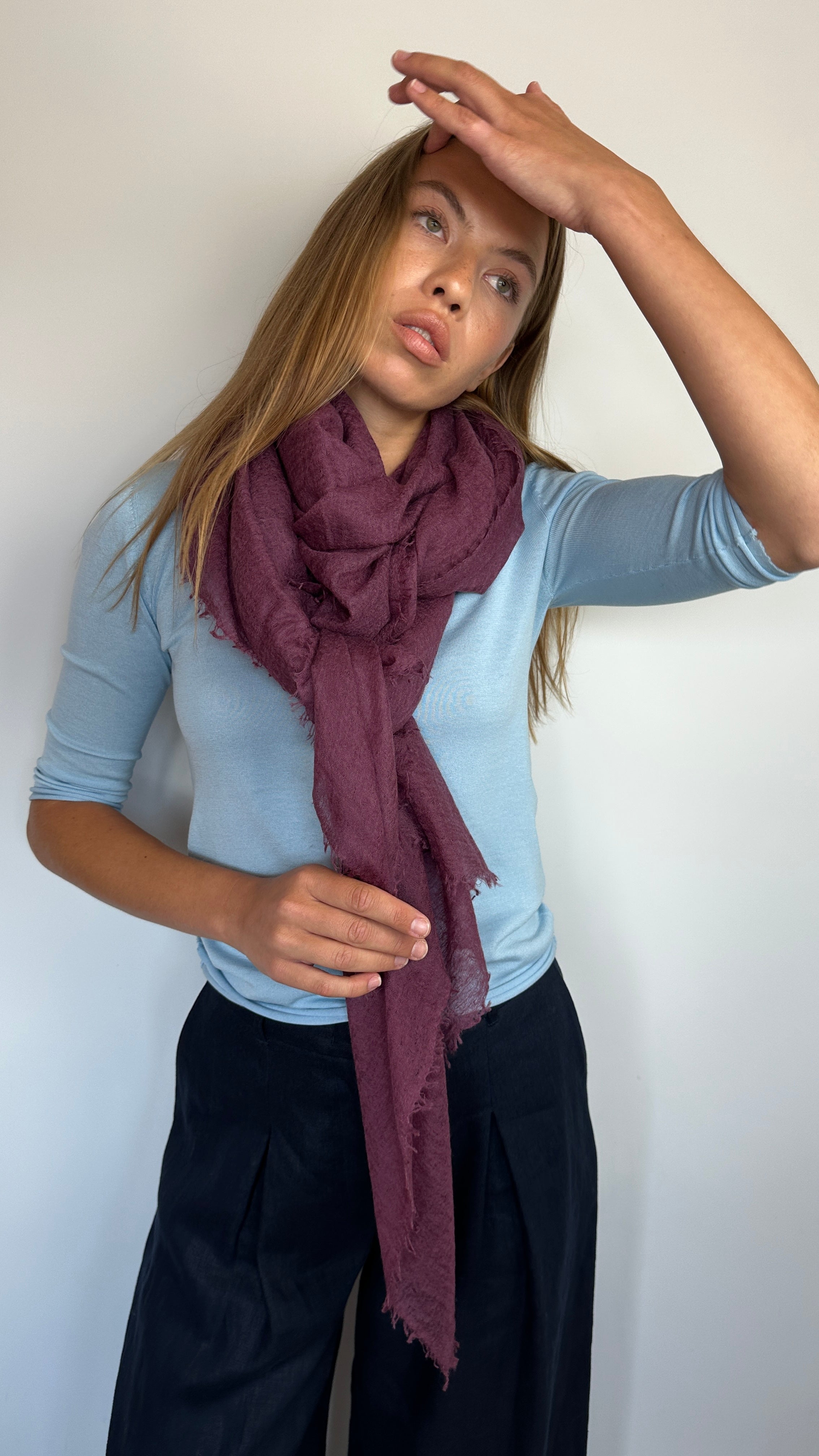 A woman wearing a light blue long-sleeve top, black pants, and a dark plum scarf, posing against a plain white wall.