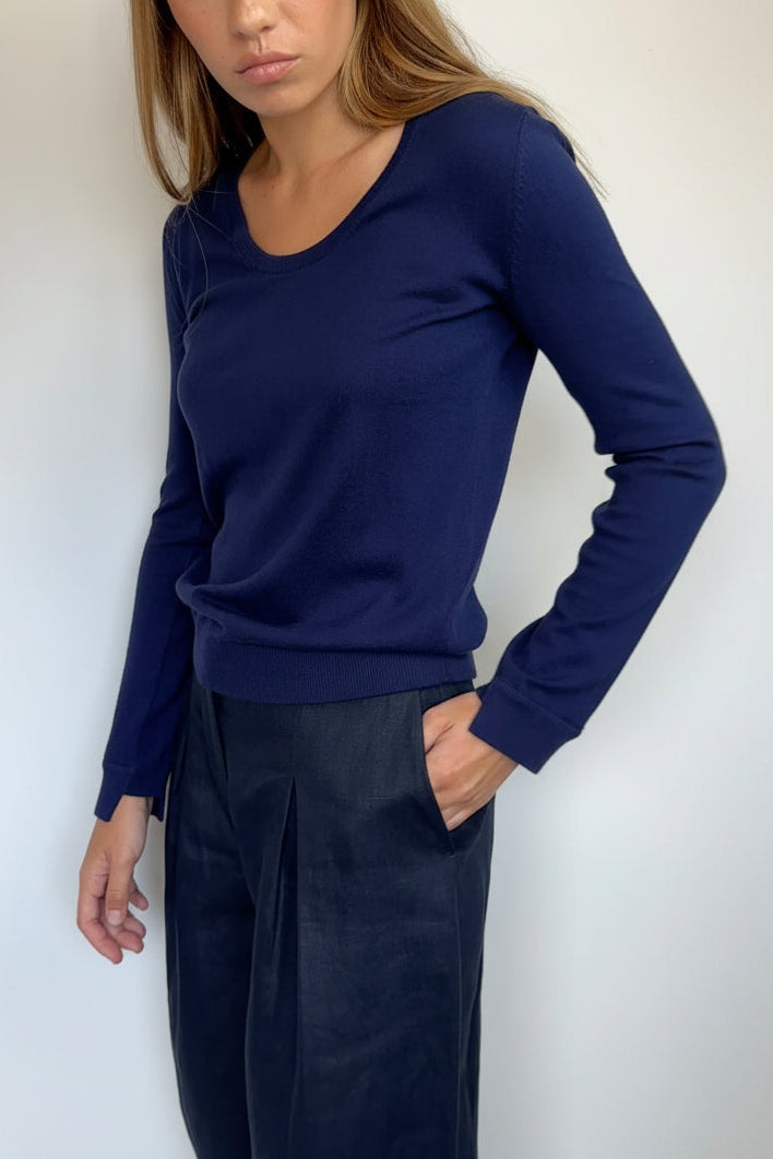 A woman wearing a navy blue long-sleeve knit top and dark navy wide-leg trousers, standing against a plain off-white wall.