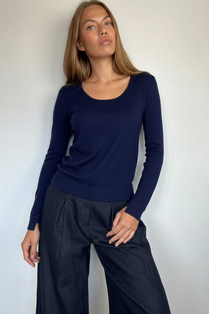 A woman modeling a dark navy blue long-sleeve scoop-neck knit top paired with dark wide-leg trousers, standing against a plain light gray wall.