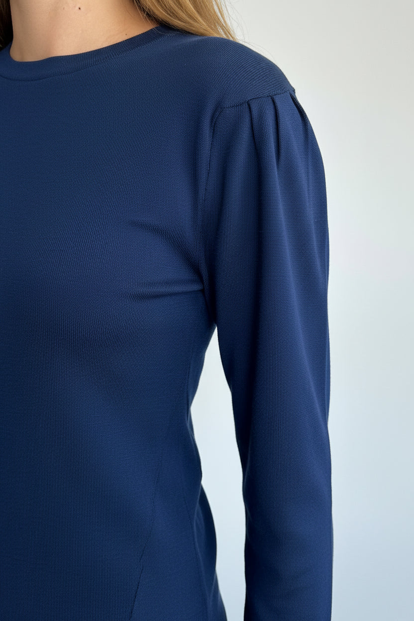 A woman with long blonde hair wearing a dark navy blue long-sleeve top with puffed shoulders, shown in a side profile against a plain light gray background.