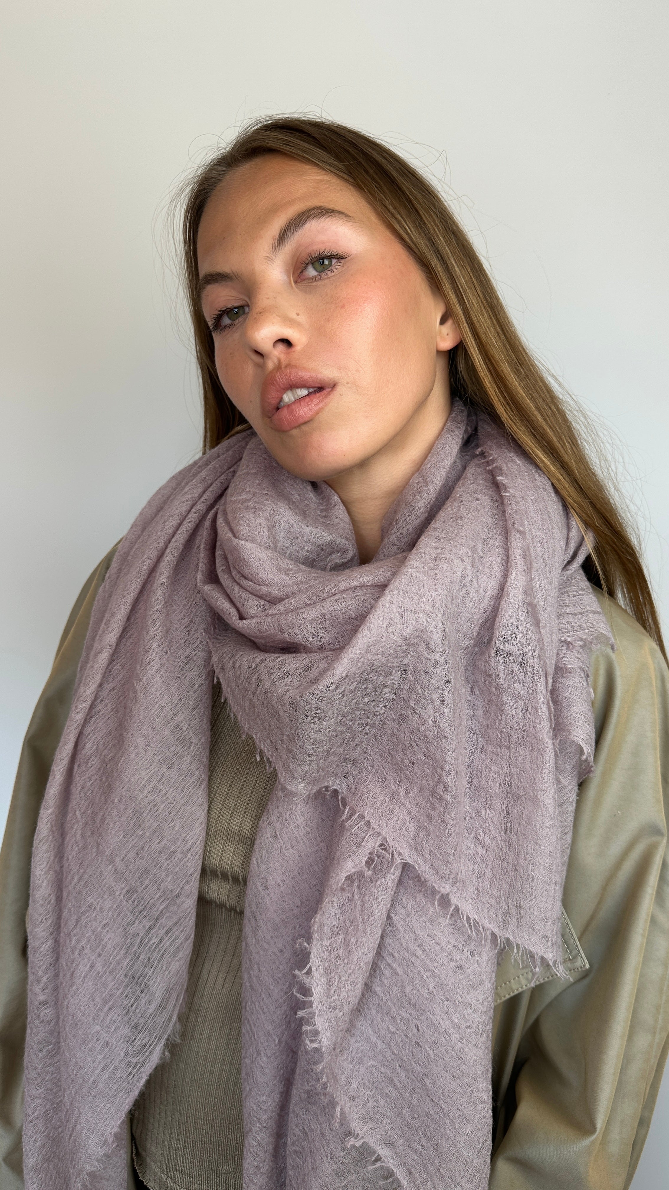 A woman with long light brown hair is shown from the chest up, wearing a soft, textured dusty mauve scarf wrapped around her neck over a beige top, against a plain off-white background.