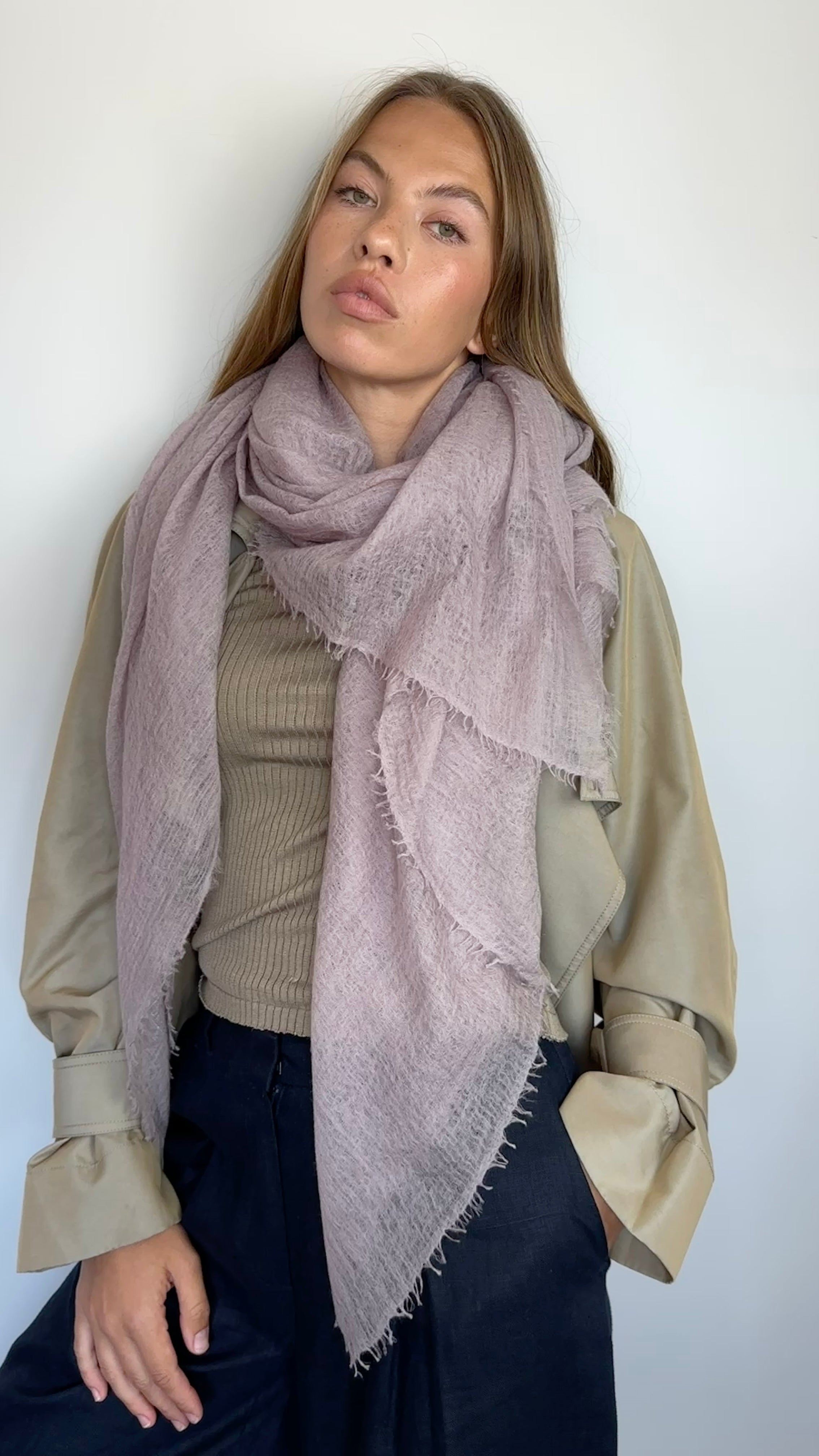 A woman modeling a large, dusty mauve-pink fringed scarf draped over her shoulders, paired with a beige top and dark navy pants against a plain white wall.