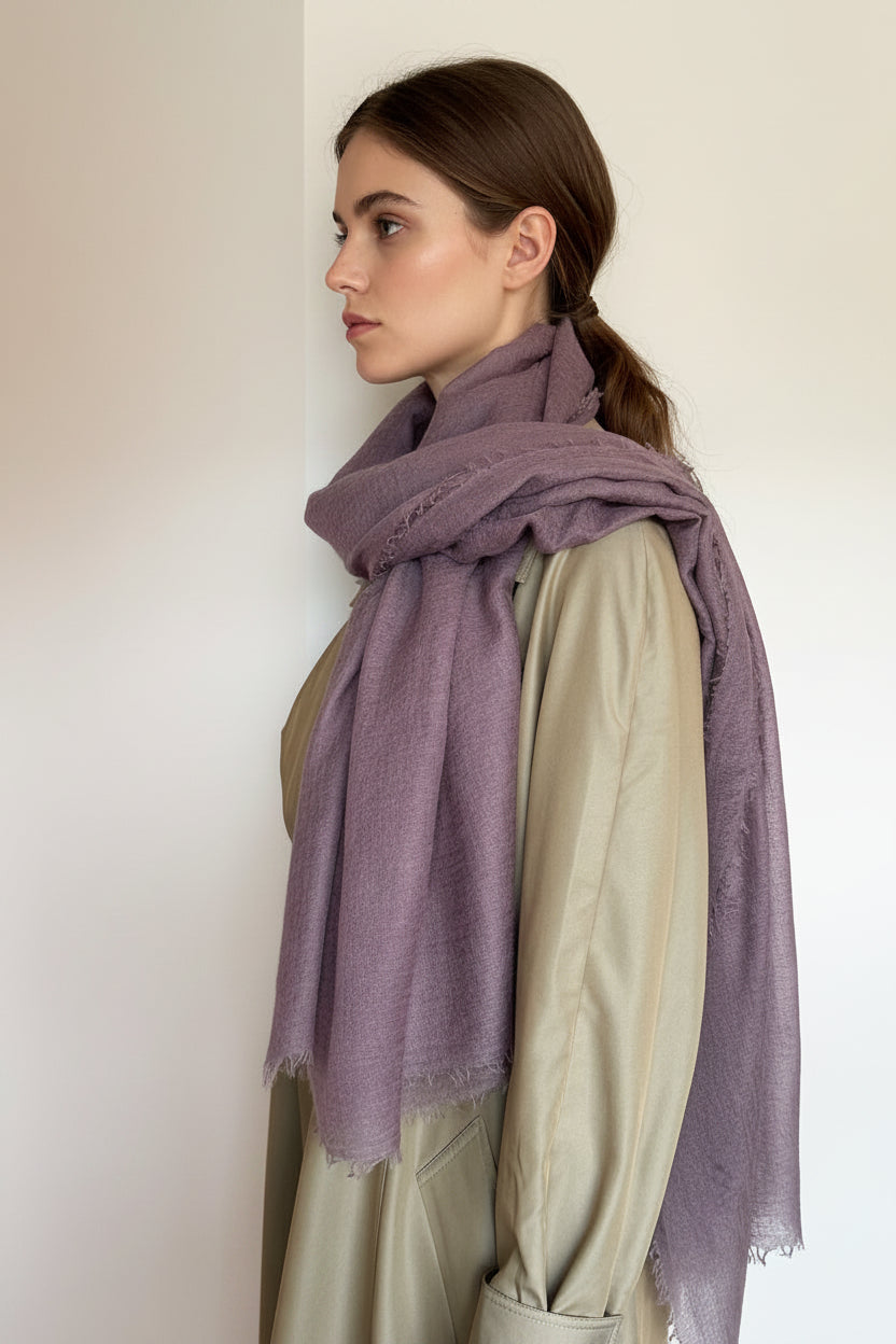 A mauve-purple scarf with frayed edges draped over a beige trench coat on a model set against a plain off-white wall.