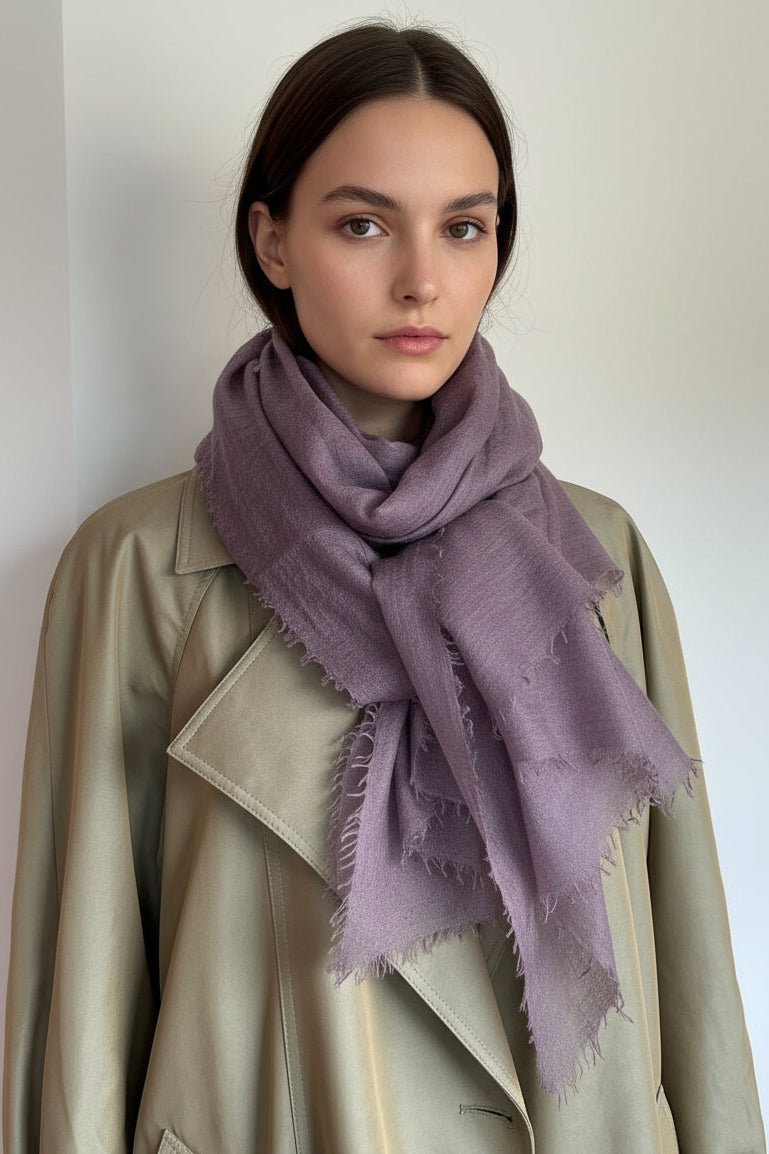 A mauve-purple scarf with frayed edges draped around a model wearing a beige coat, set against a plain off-white wall.