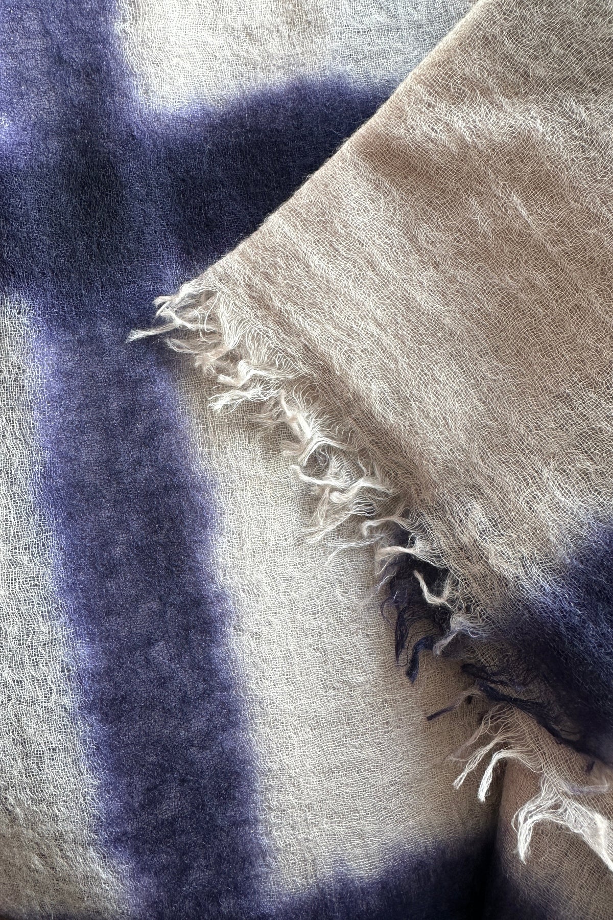 A close-up of a soft woven textile with a large cross-like pattern in deep purple over an off-white base, with frayed fringe visible along one edge.