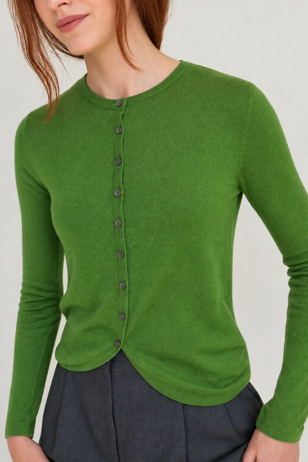 A woman wearing a bright green long-sleeve knit top with a buttoned front and dark gray trousers, shown from the neck down against a plain light background.
